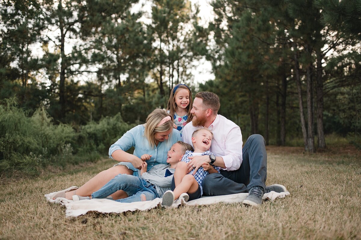 Family of 5 sitting on blanket in the grass tickling each other and laughing by Cypress Texas Photographer