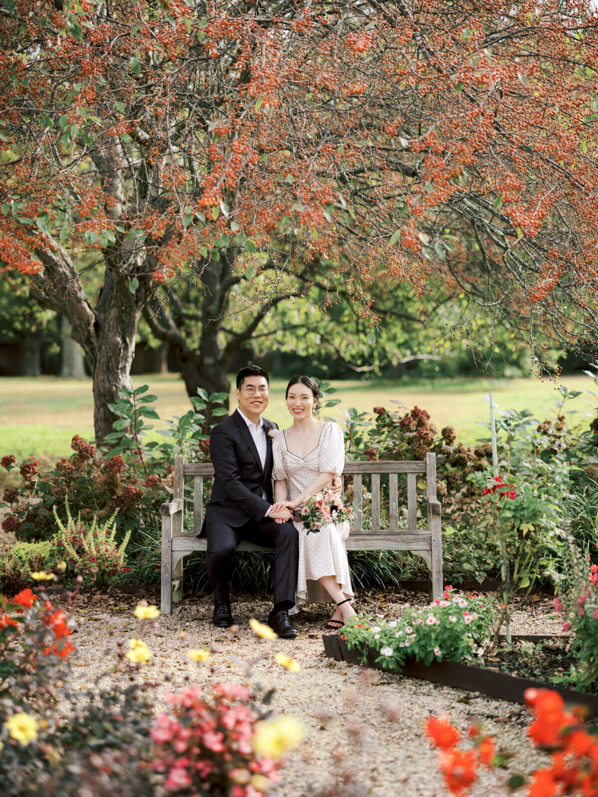 The engaged couple is sitting on a wooden bench amidst beautiful foliage in orange and yellow hues. Image by Jenny Fu Studio