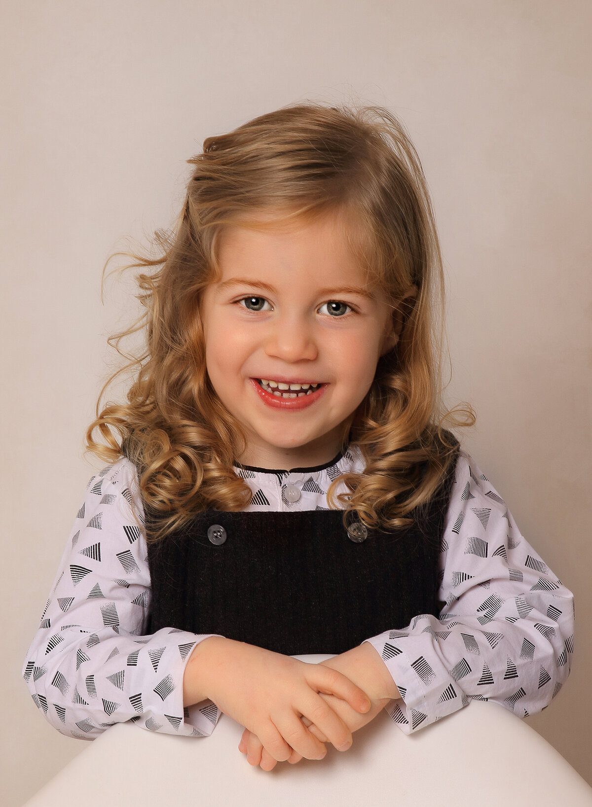 Little girl is sitting for a portrait in a brooklyn photo studio. She has her hans folded on a table in front of her and is smiling at the camera with curly blonde hair. Captured by Chaya Bornstein, Brooklyn Family Photographer