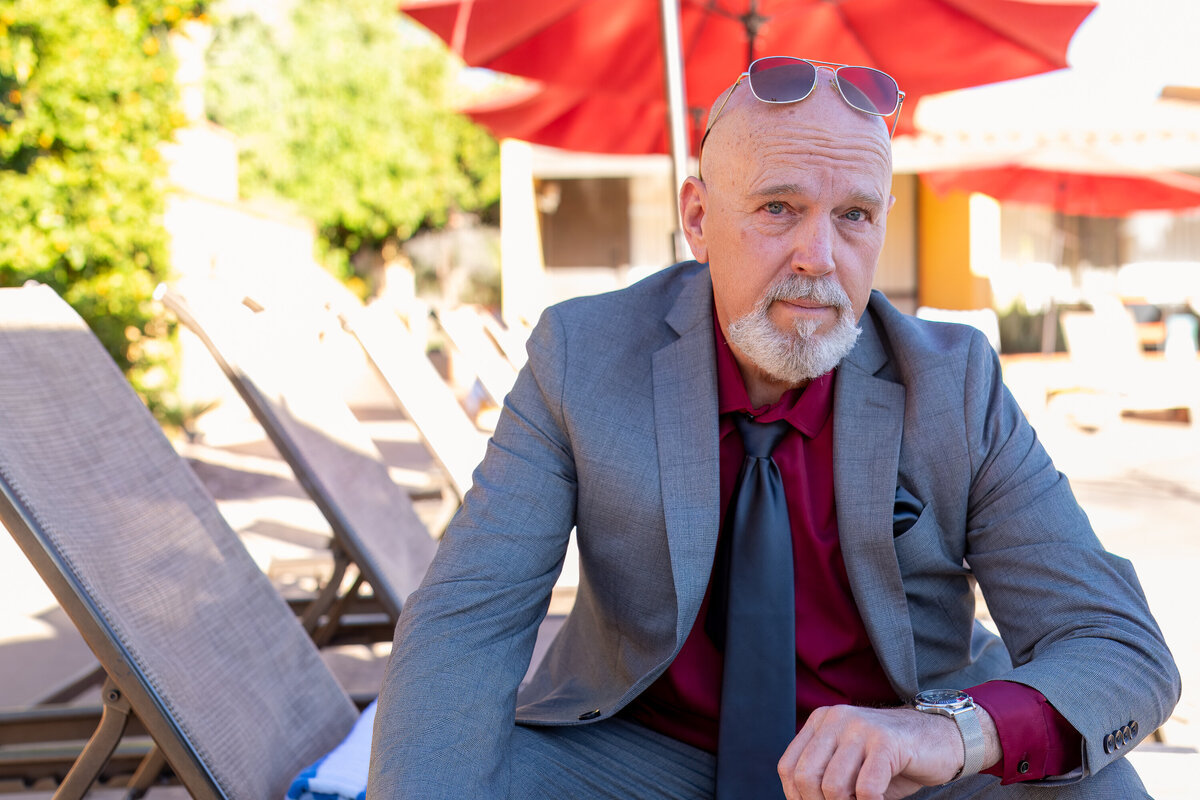 Man in a blue suit and red tie sitting outdoors near poolside lounge chairs and red umbrellas, photographed by Vyrl Photo for Tucson brand photography.