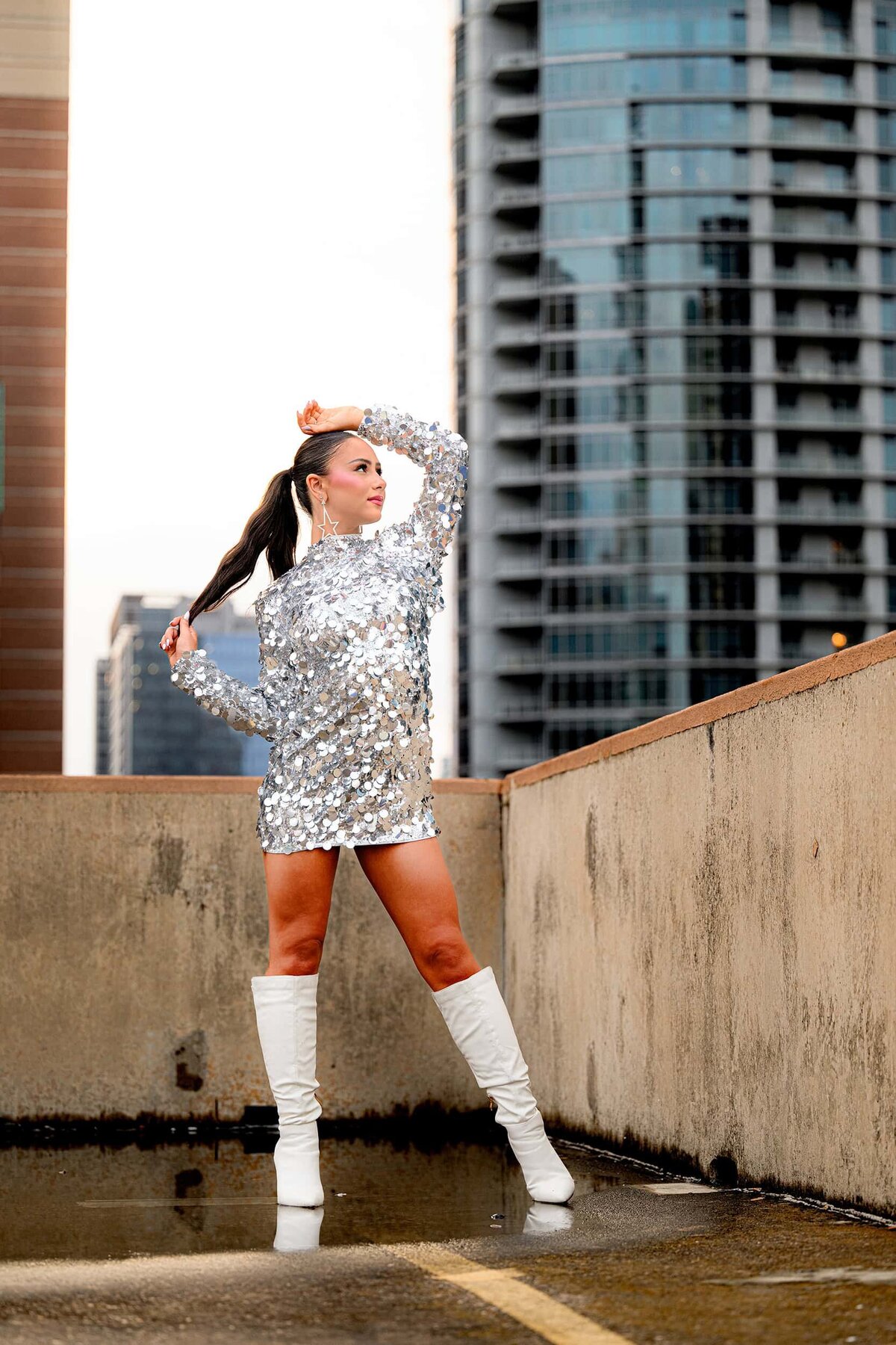Senior girls stands on a rooftop in a silver sequined dress wearing white boots and holding her ponytail in her right hand and her left hand on her head during senior portraits
