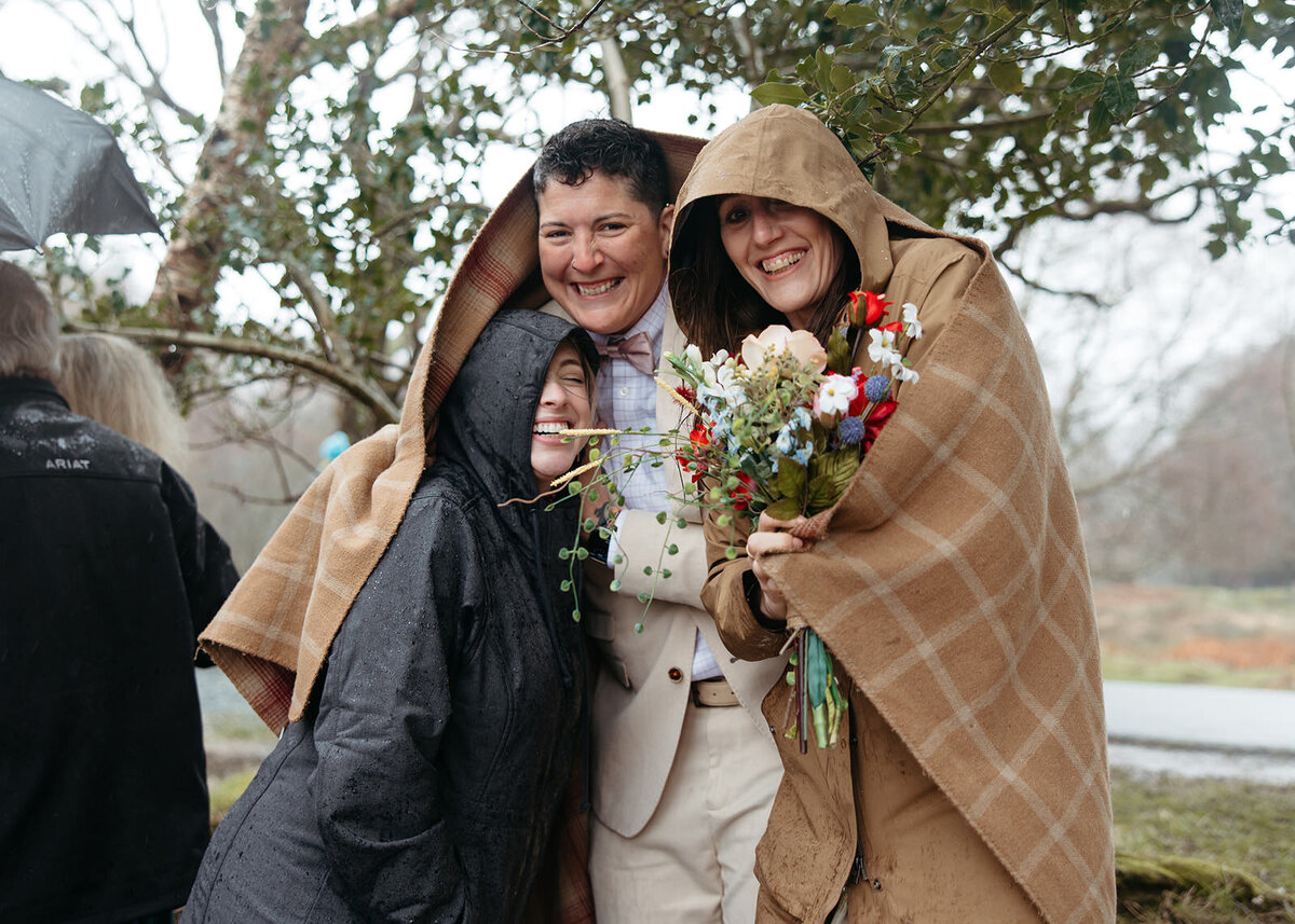 Three people huddled under a plaid blanket, smiling with a bouquet of flowers in a rainy outdoor setting