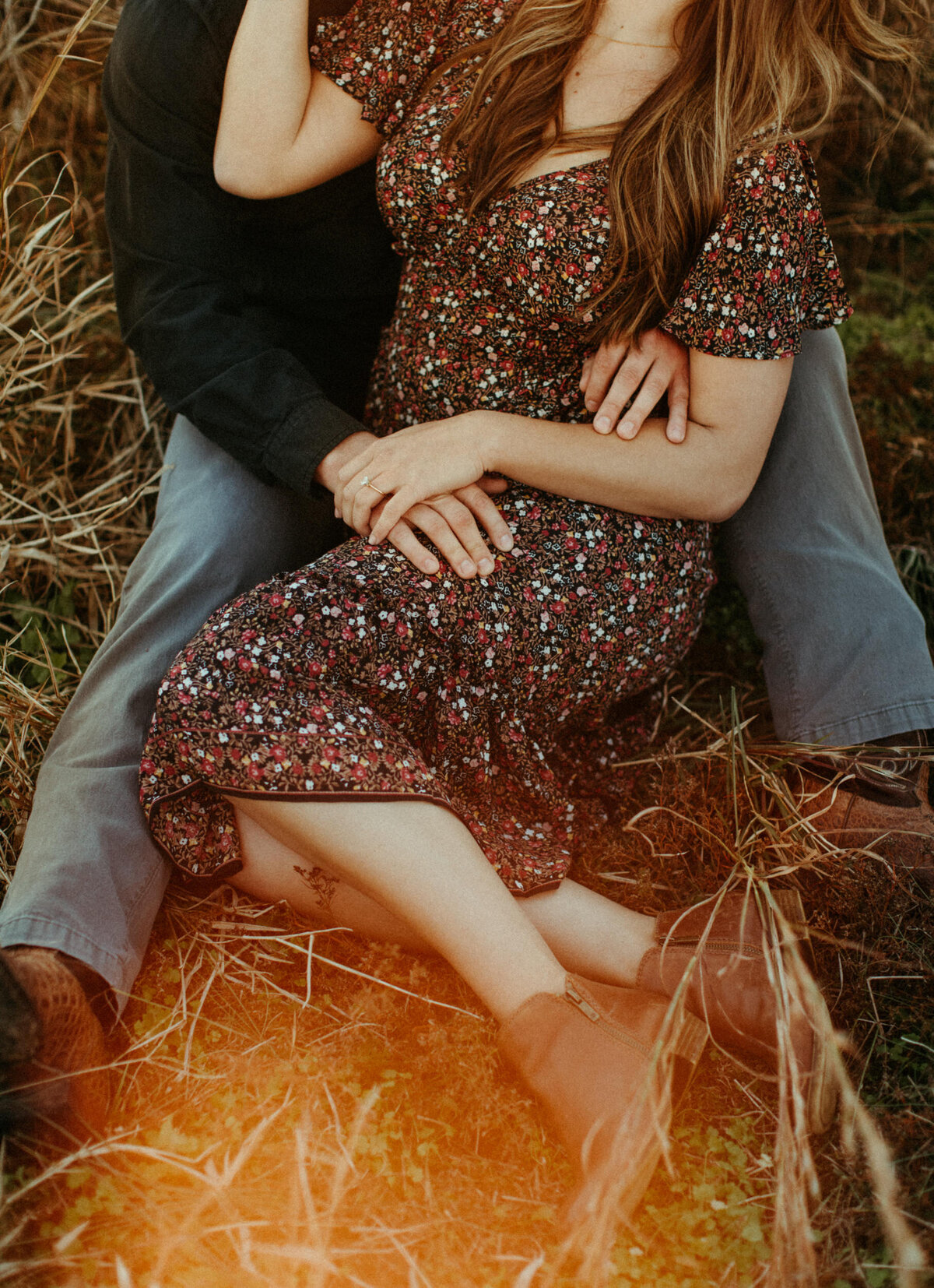 A girl in a dress is sitting on the ground between her fiancé's legs while he wraps his arms around her.