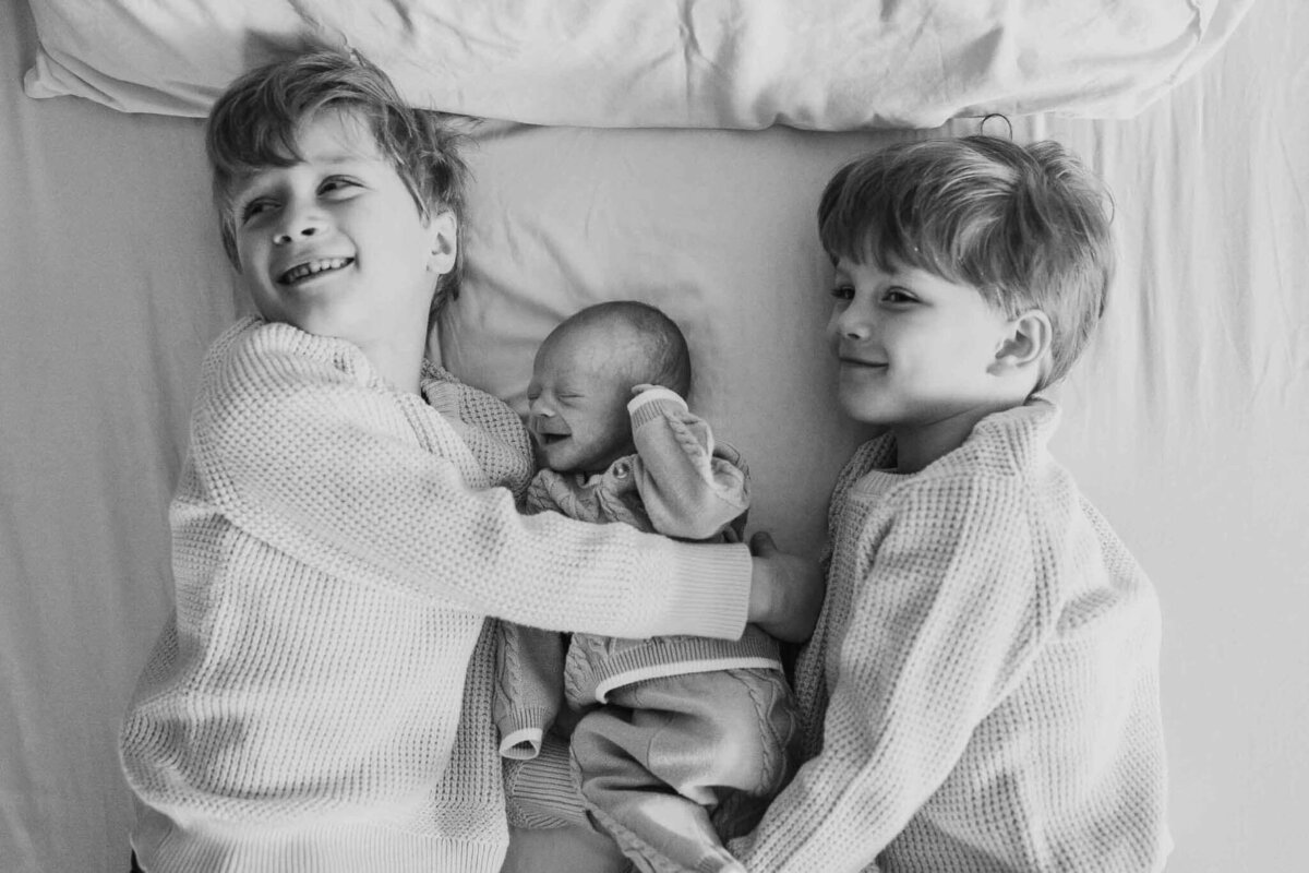 Two big brothers cuddle their newborn sibling on a soft bed, sharing warm smiles during an in-home lifestyle session.