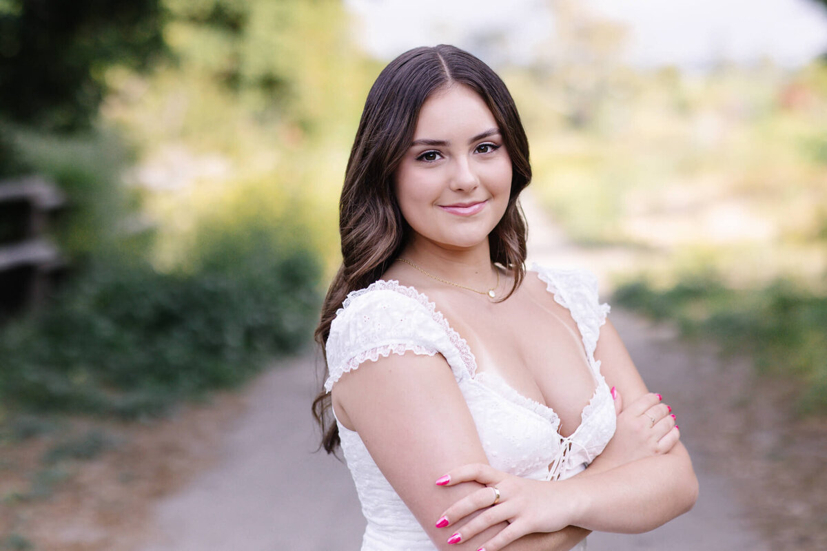 A woman with long dark hair, wearing a white lace dress, stands outdoors with her arms crossed. She is smiling, with a blurred green and natural background behind her.