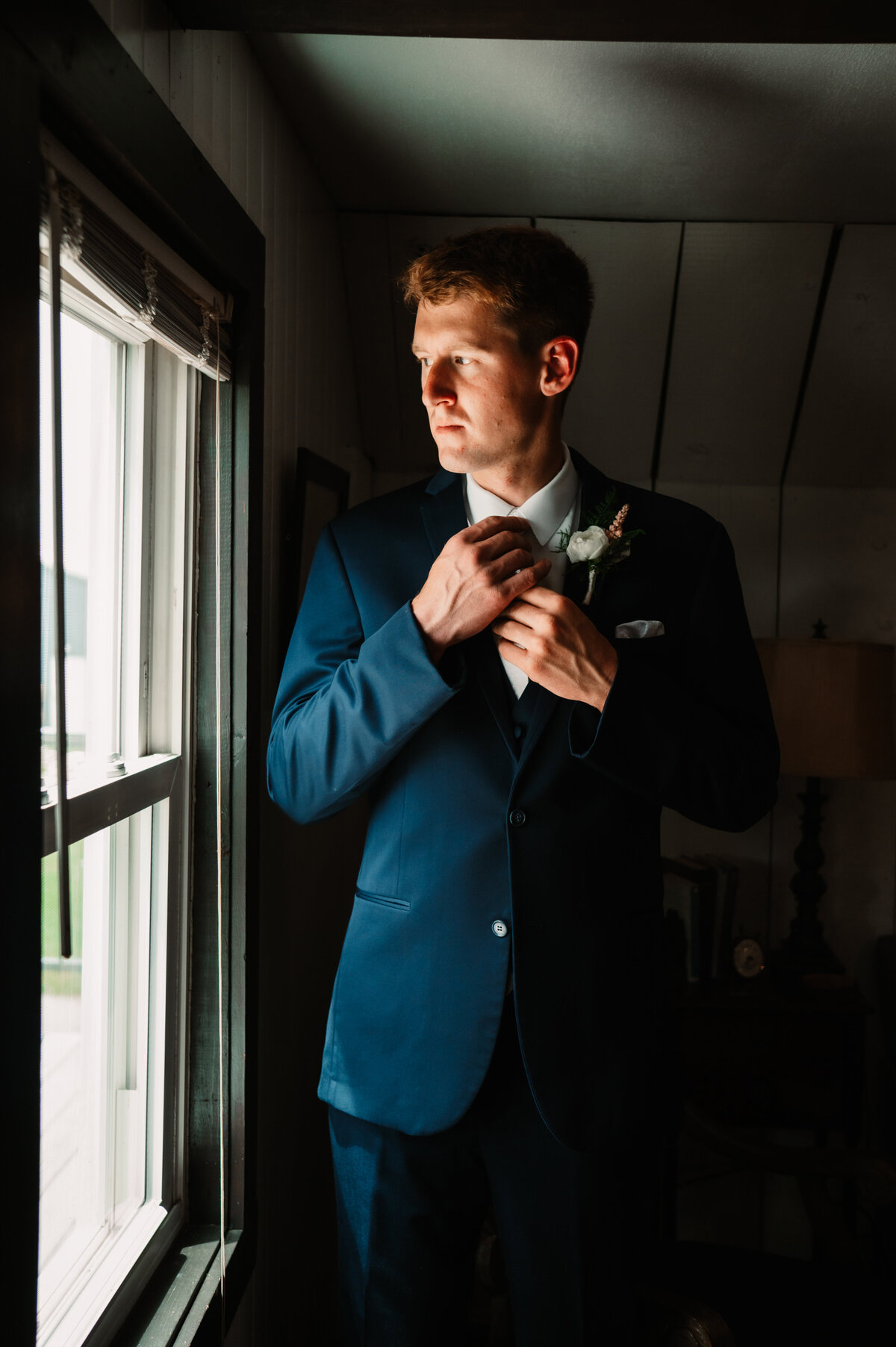 A groom in a blue suit adjusts his tie in a window