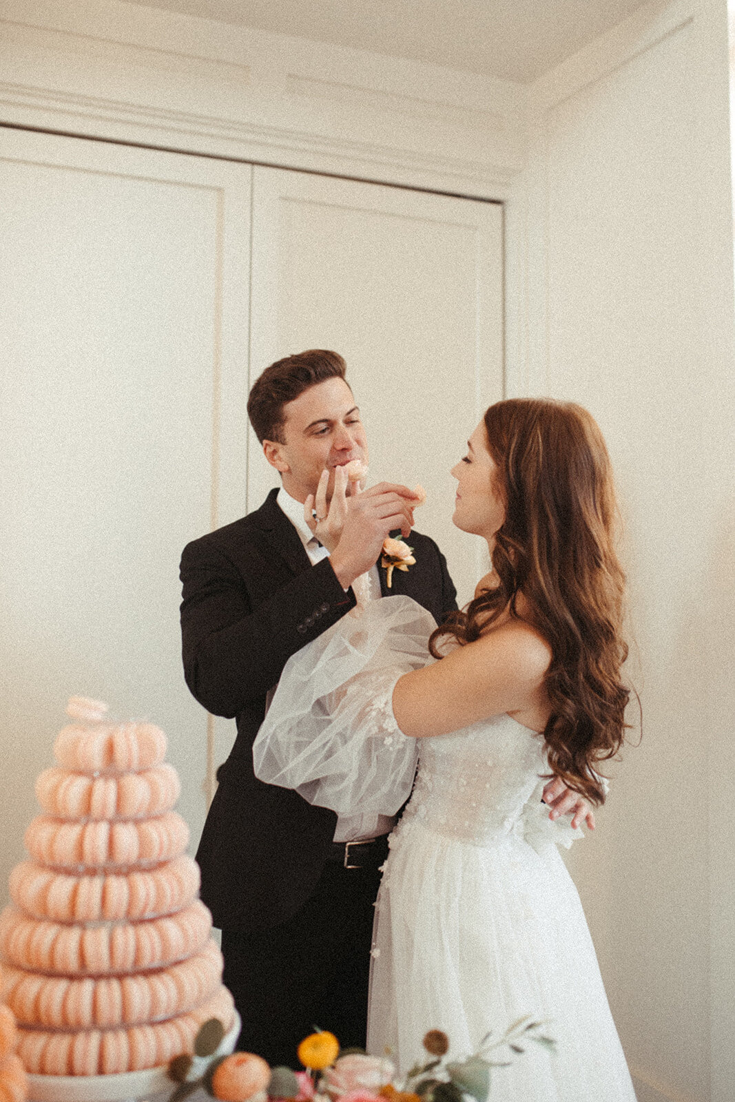 A bride and groom wearing a white wedding gown and black tuxedo feed macaroons to each other next to the dessert table.