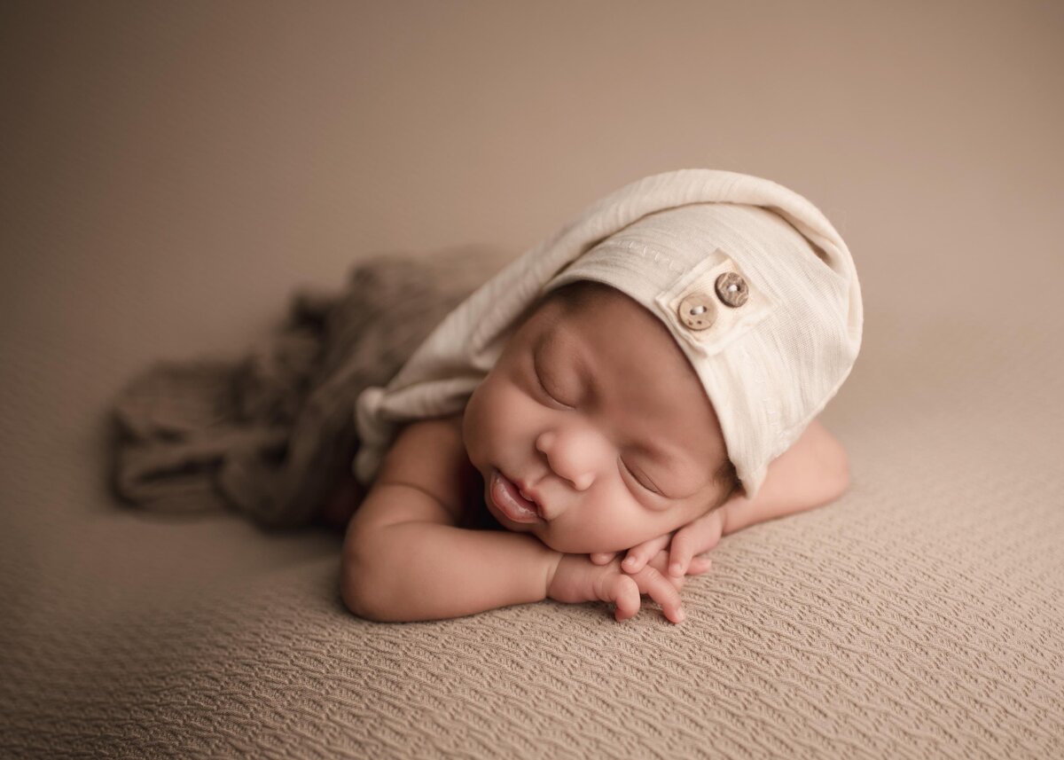 Baby boy is posed for his Lake Elsinore newborn photoshoot. He is laying on his belly wearing a long nightcap with button details. His hands are folded underneath his cheek and he is sleeping. Captured by best Lake Elsinore newborn photographer Bonny Lynn Photography.