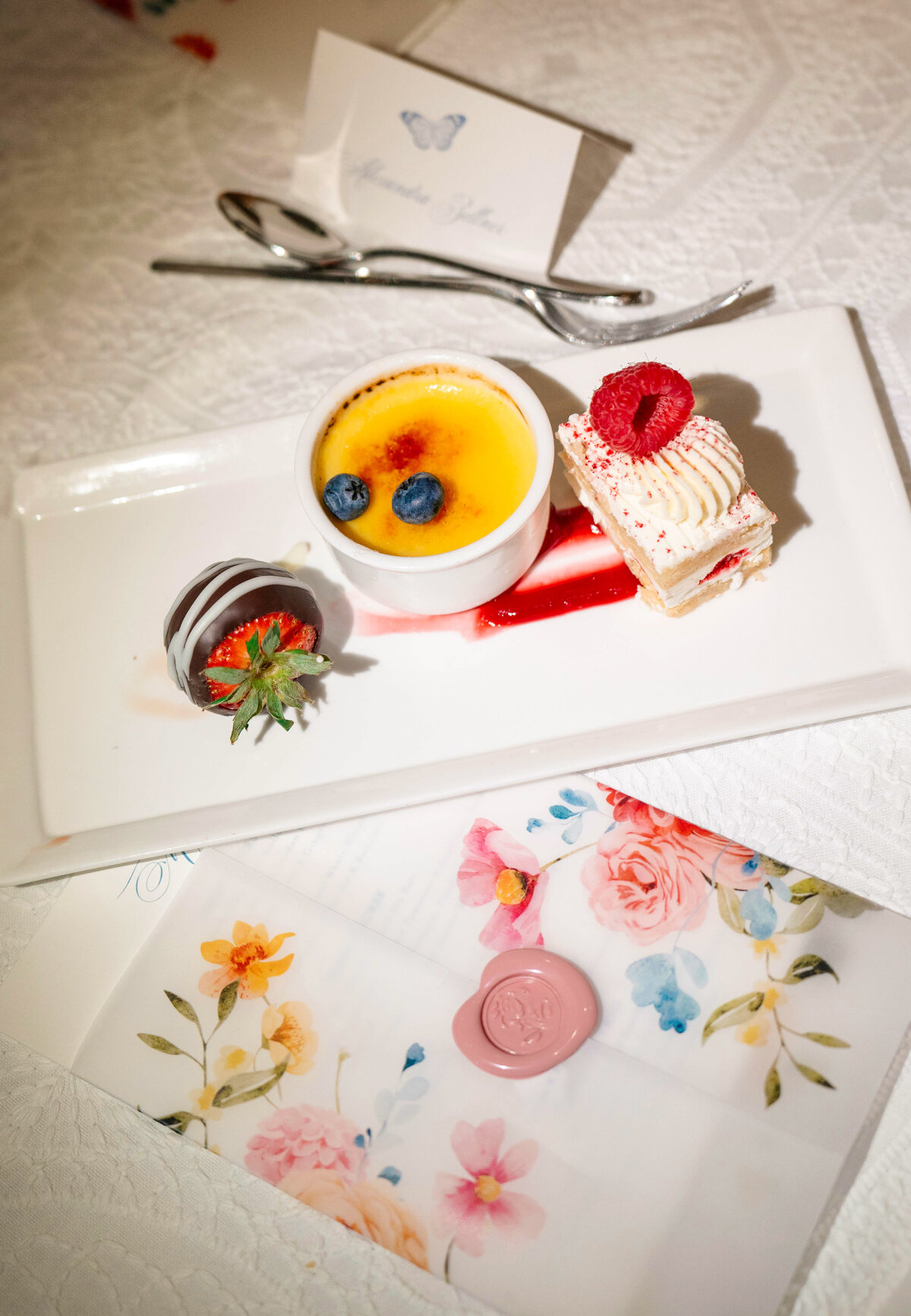 A rectangular white plate with a creme brulee topped with blueberries, a chocolate-covered strawberry, and a piece of cake garnished with a raspberry. Nearby, floral stationery with a wax seal and cutlery on a white tablecloth.