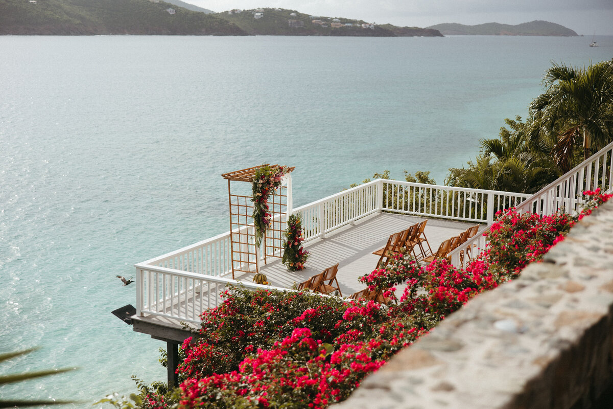 A scenic oceanfront wedding setup with wooden chairs and bright pink bougainvillea flowers overlooking a tranquil bay