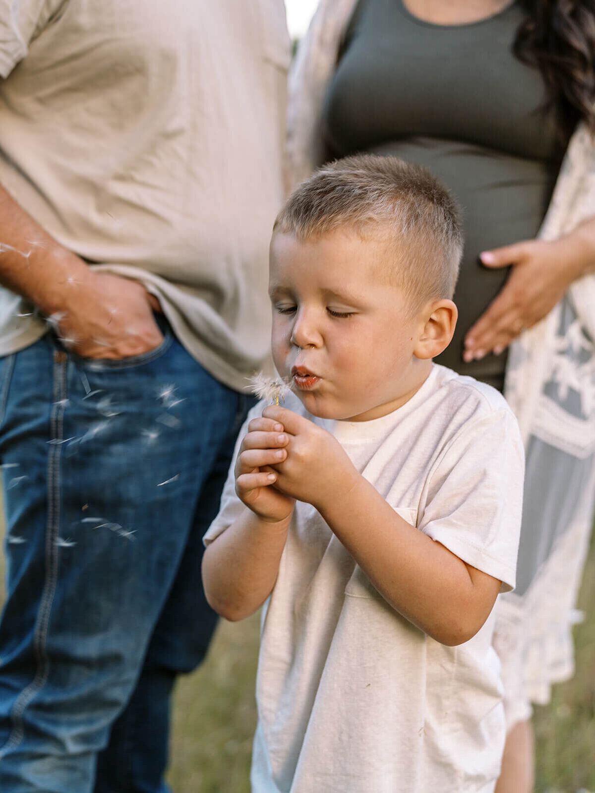 A young boy blows on a dandelion with his parents in the background.