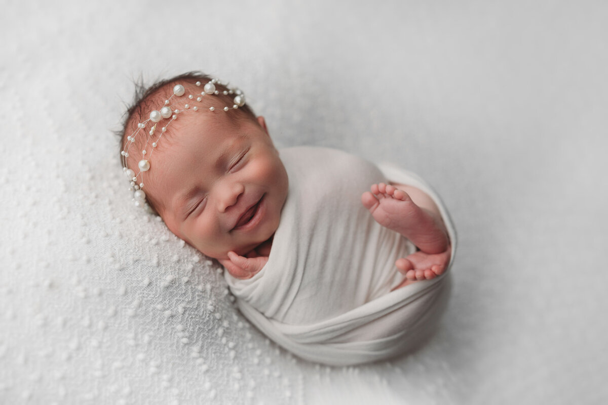newborn baby girl smiling with pearl headband white wrap on all white backdrop with feet poking out