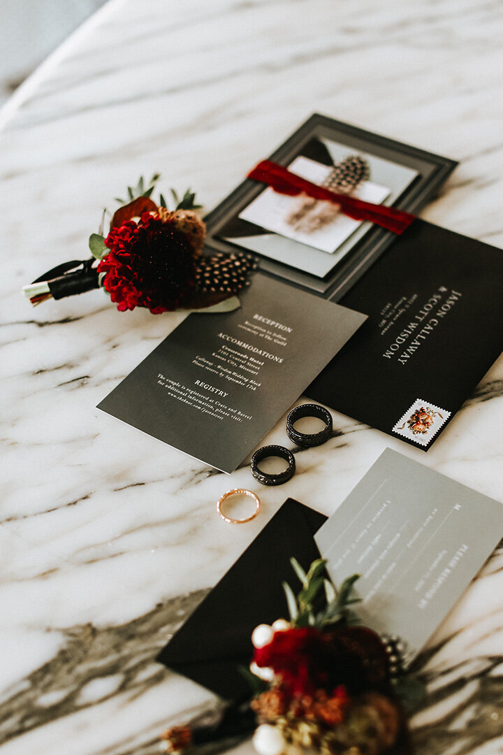 Mixed wedding stationery with white font on a marble counter with wedding bands and flowers.