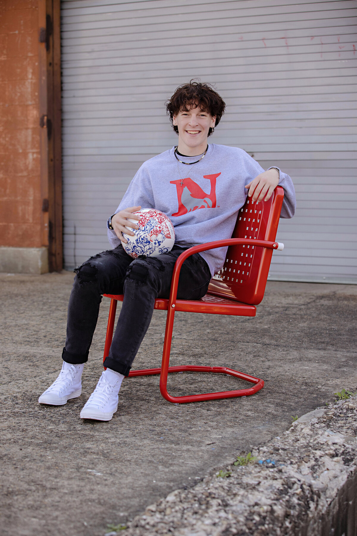 High school senior seated on a red metal chair holding a soccer ball and wearing a college sweatshirt, with metal loading dock bay doors behind, photographed in Charlotte NC.