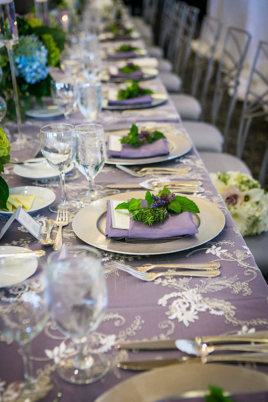 place setting with silver charger plates, puple napkins, lace linens, and greenery on each napkin