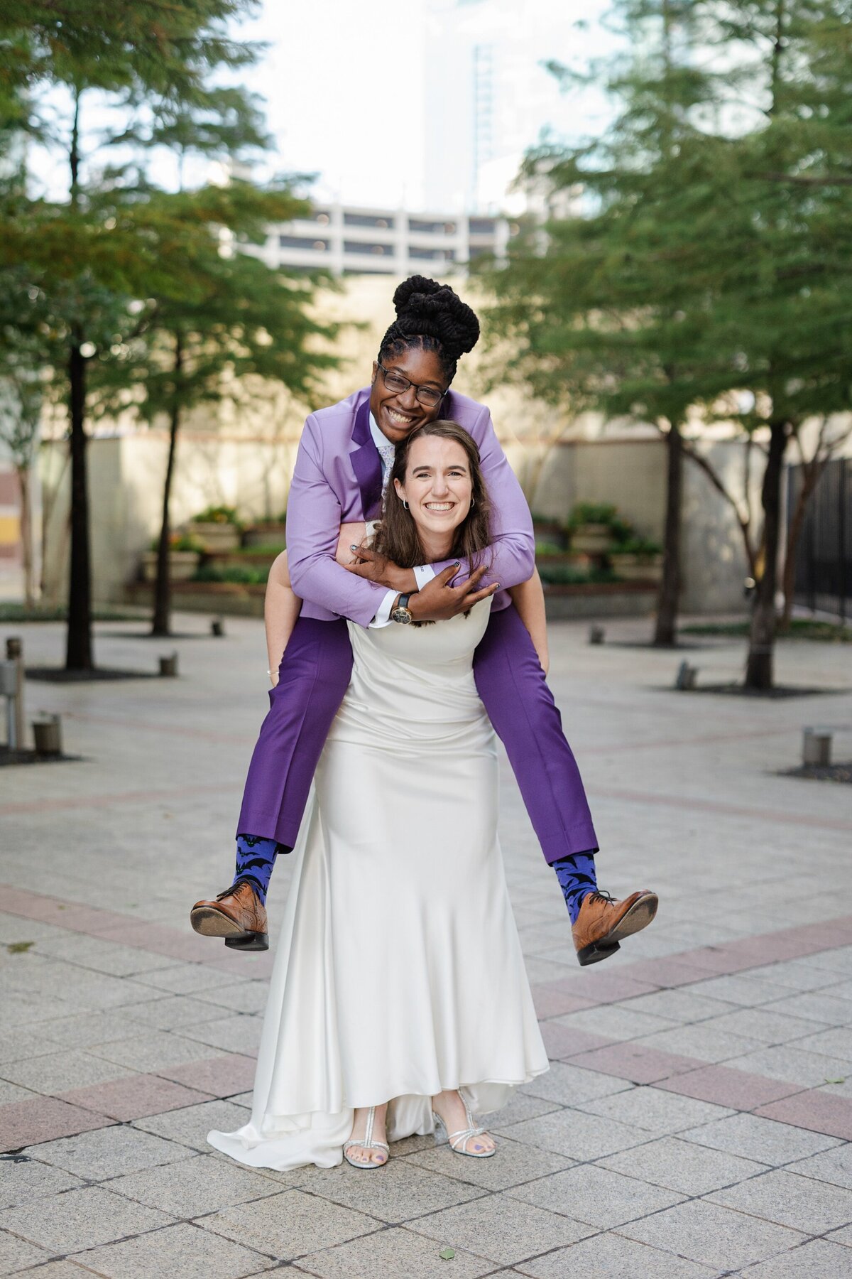 A playful portrait of a two brides on their wedding day at The Westin Dallas Downtown in Dallas, Texas. One bride is riding "piggyback" on the other and both are joyfully smiling at the camera. The bride on the bottom is wearing an elegant white dress and heels while the bride being held up is wearing a purple suit with brown dress shoes.