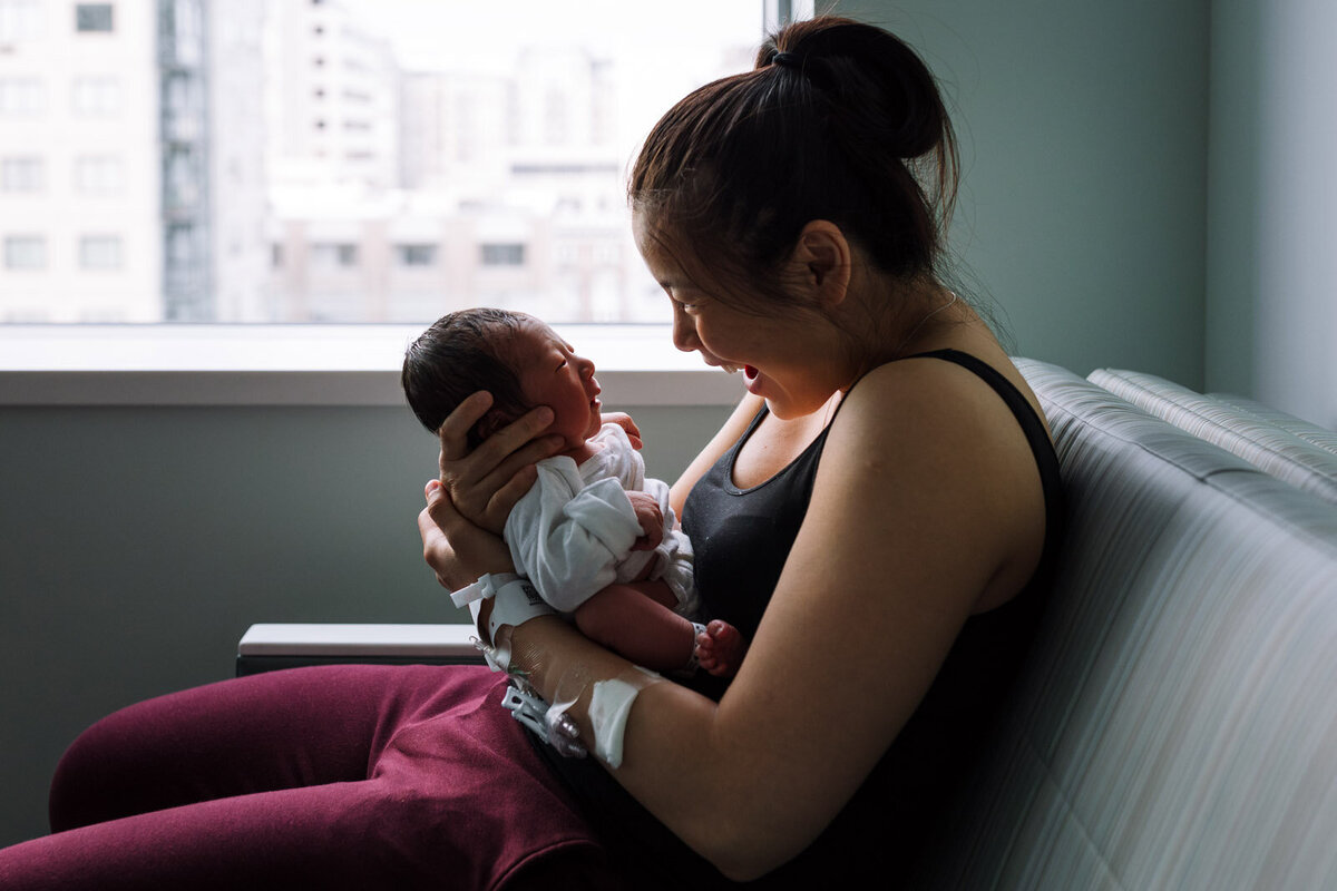 mom sitting on hospital couch holding newborn close to her face and smiling as he gazes at her