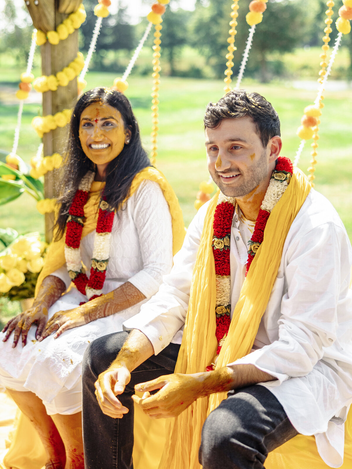 A couple dressed in white attire with yellow scarves and floral garlands sits smiling amidst a sunny outdoor setting. Both have turmeric paste on their faces, celebrating a joyful occasion with marigold decorations around them.
