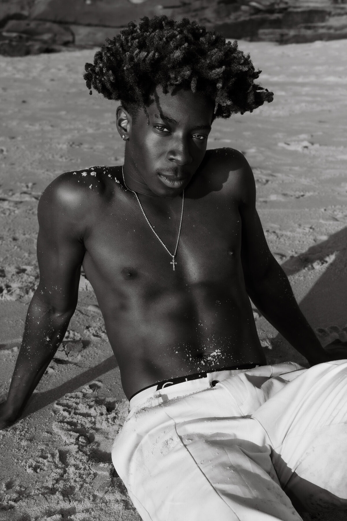 black and white headshot of a man wearing white linen pants and a gold cross chain necklace with white sand placed on his arms and chest