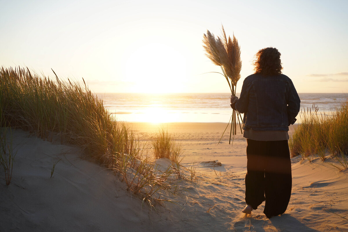 Woman  carrying pampas grass, walking  on a sandy  dune toward the ocean at sunset