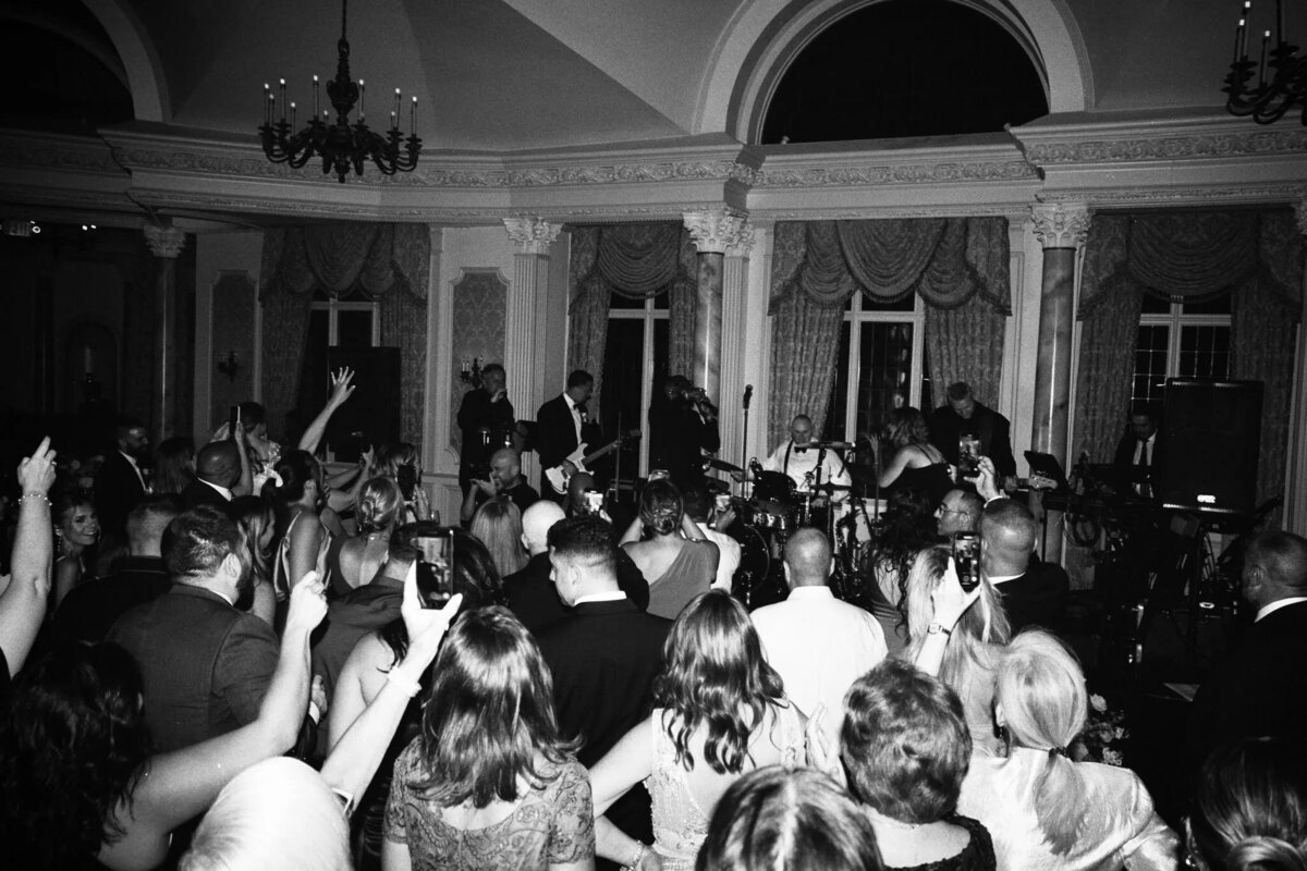 A black-and-white photo by a film photographer NJ captures a crowd at an indoor concert, everyone facing the band on stage, with raised hands and phones held high to snap pictures or record video.