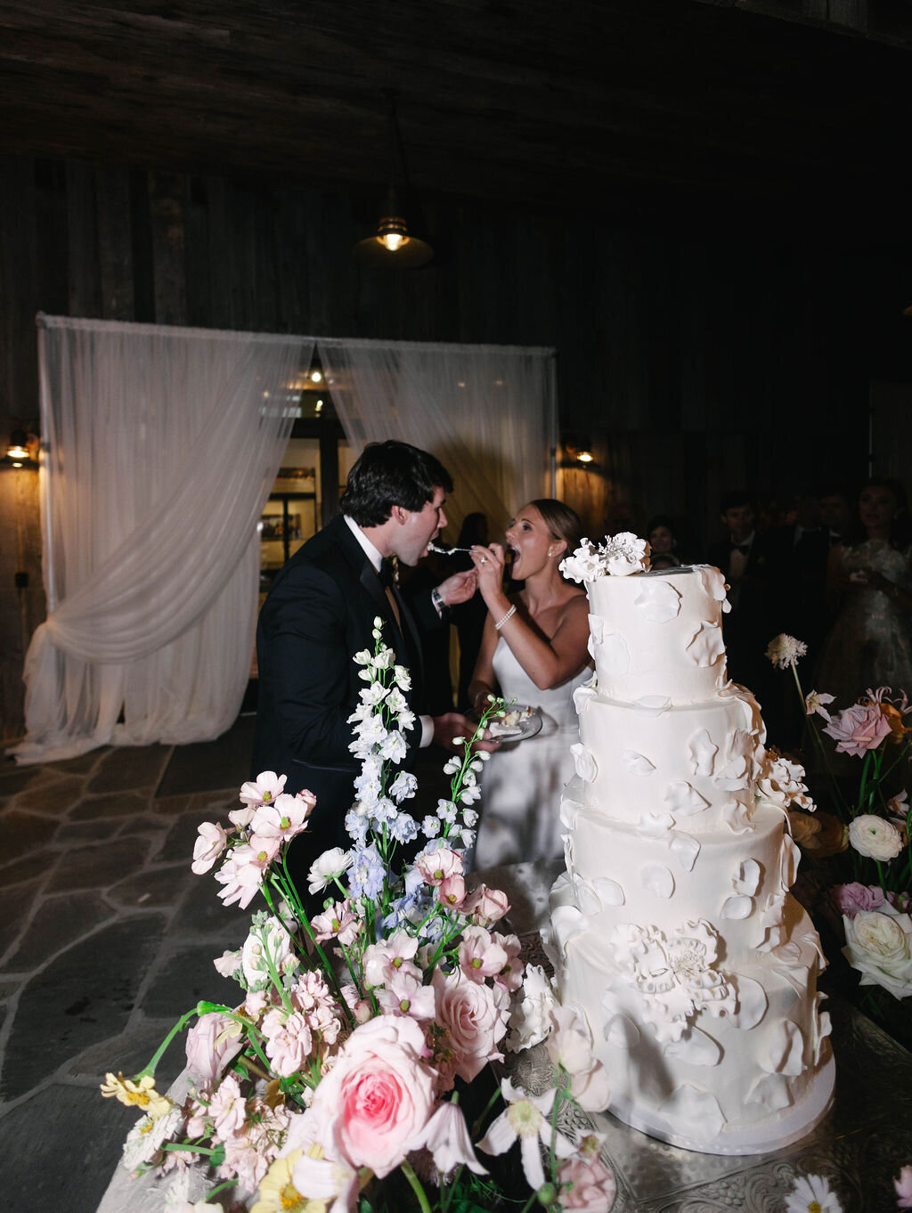 Bride and groom feed each other cake during their reception at The Bascom in Highlands, NC, with floral décor and a four-tier white wedding cake.