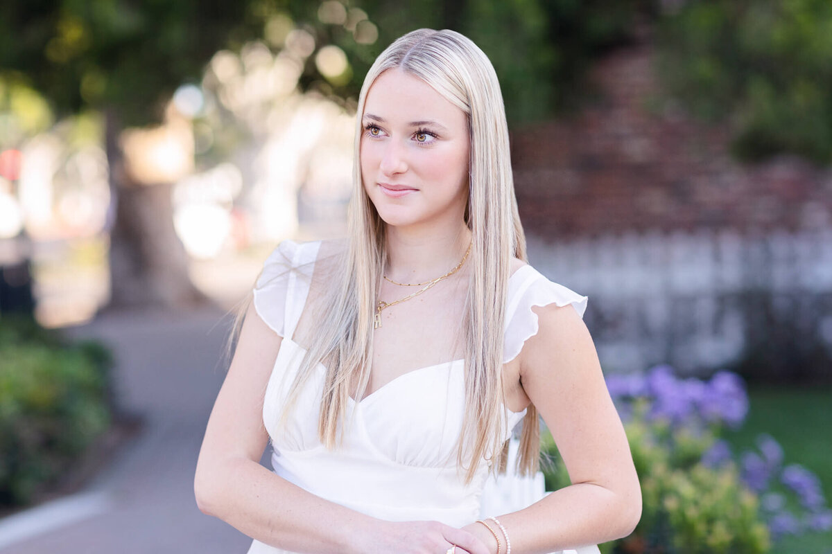 A woman with long blonde hair stands outdoors, wearing a white dress, looking to the side. The background is blurred, with hints of greenery and flowers.