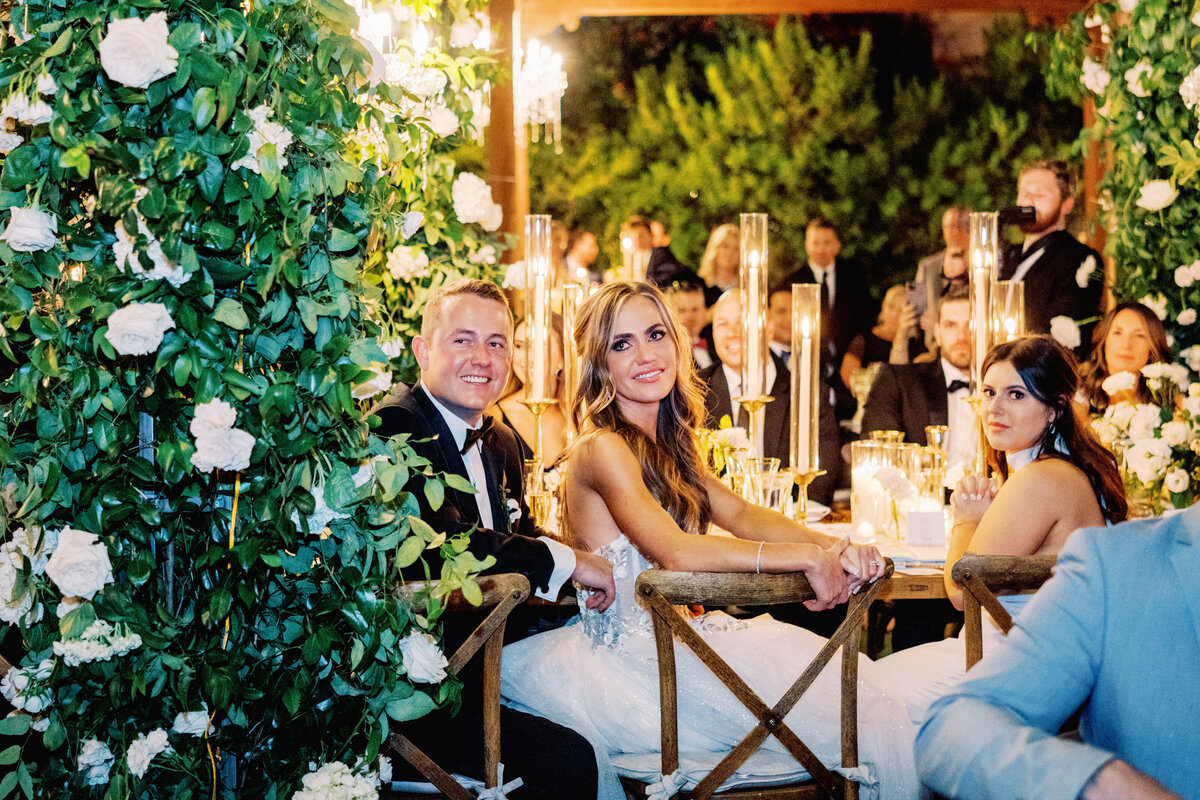 The newlyweds enjoy a romantic moment at their candlelit reception table, surrounded by lush greenery, elegant floral decor, and flickering candlelight.