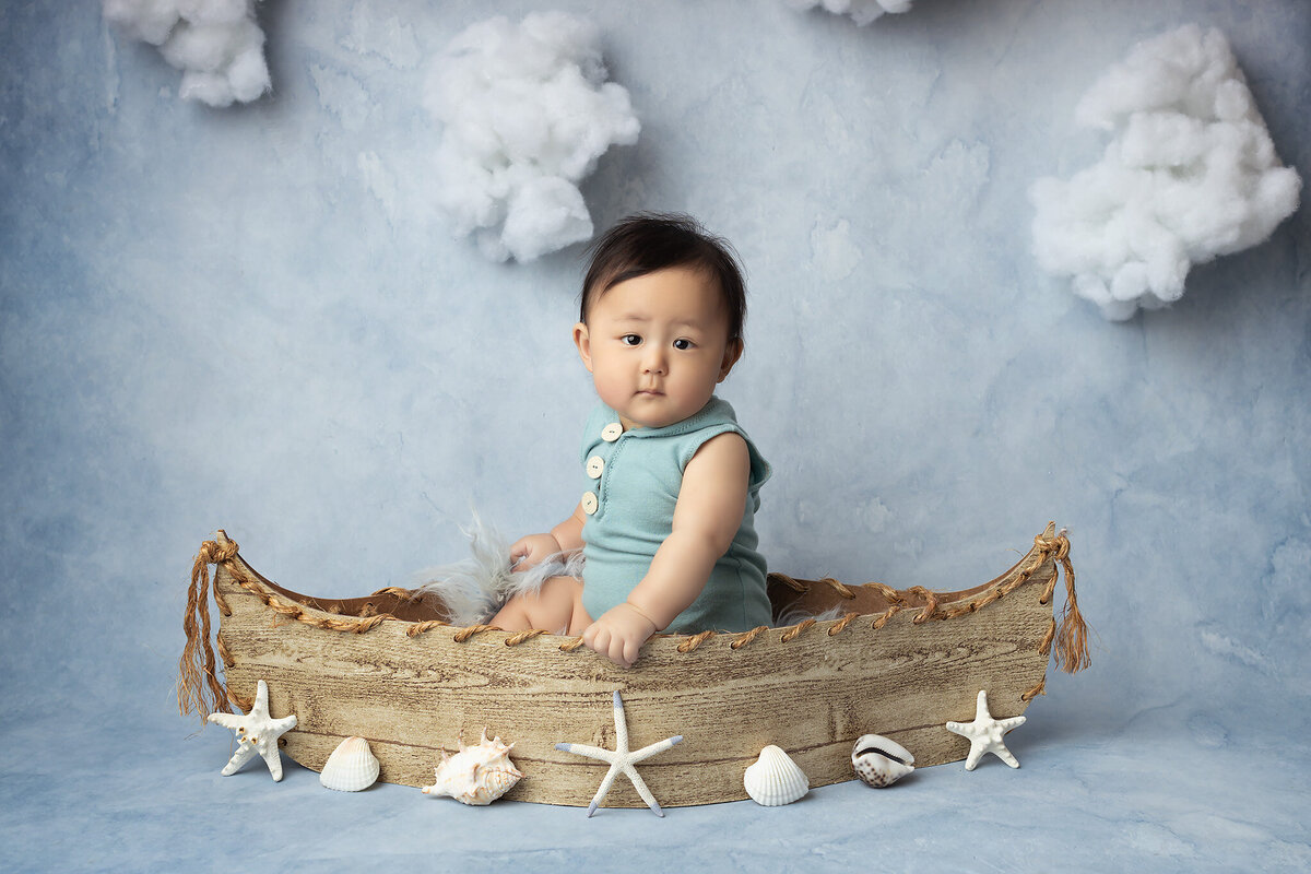 A baby dressed in a seafoam romper sits inside a wooden boat surrounded by seashells and starfish, against a dreamy backdrop of soft clouds. This whimsical image by a Dallas baby milestone photographer captures a sense of adventure and imagination.