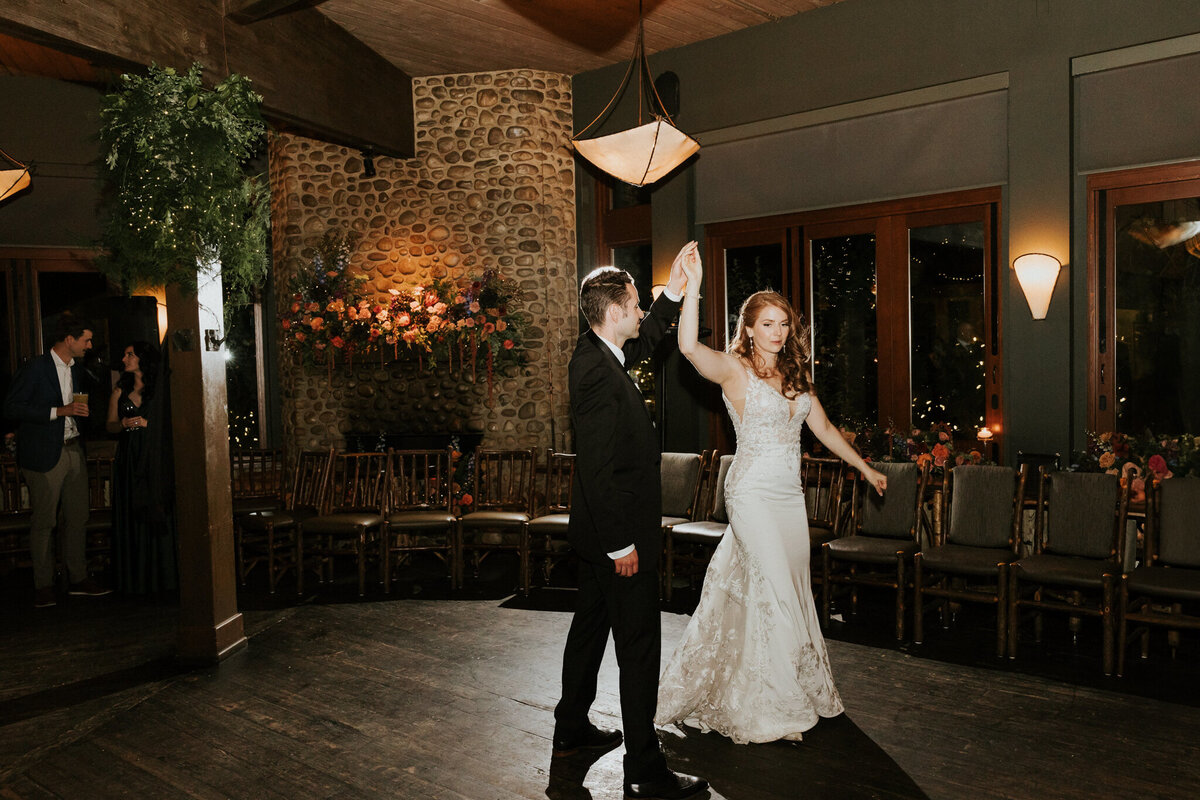 Elegant wedding reception at River Café in Calgary, Alberta, featuring the newlyweds sharing their first dance in a rustic, candlelit setting. The bride stuns in a fitted lace gown, while the groom looks sharp in a classic black suit. The cozy venue, with its stone fireplace, wooden accents, and floral arrangements, creates a warm and romantic atmosphere for this intimate celebration.