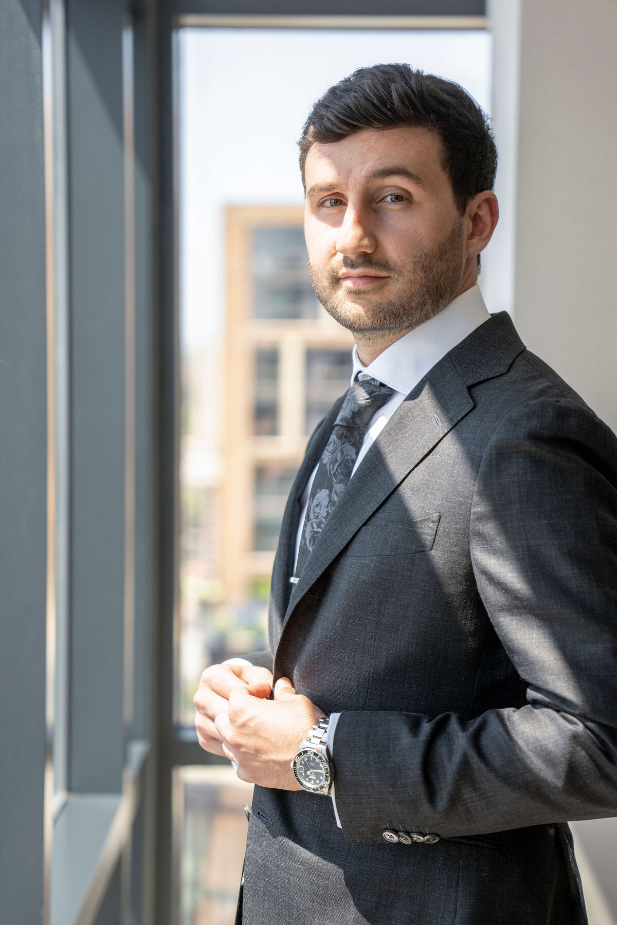 A well-dressed man in a dark suit stands by a window. He has short dark hair and a trimmed beard, wearing a watch and looking towards the camera with a confident expression. The window shows blurred building outlines in the background.