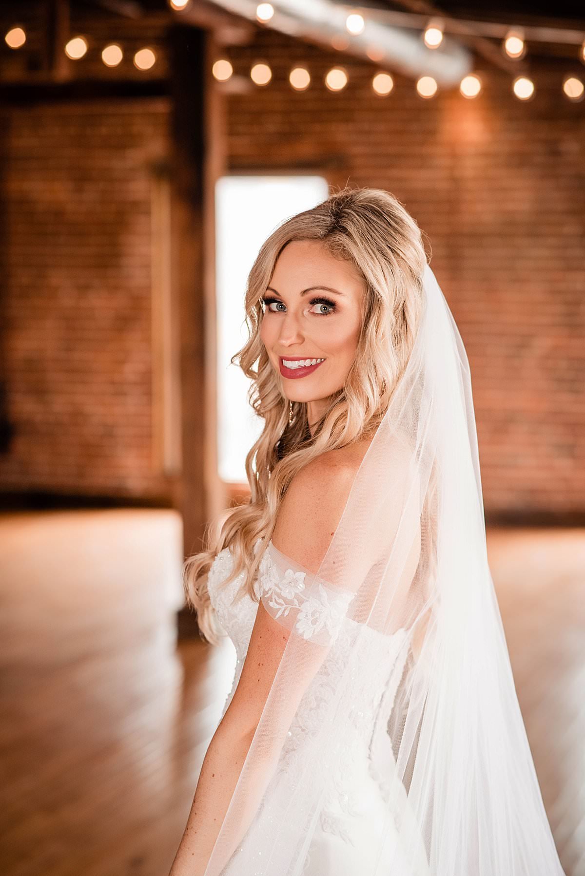 Bride looking over her shoulder at the camera, cathedral veil flowing and bistro lights above