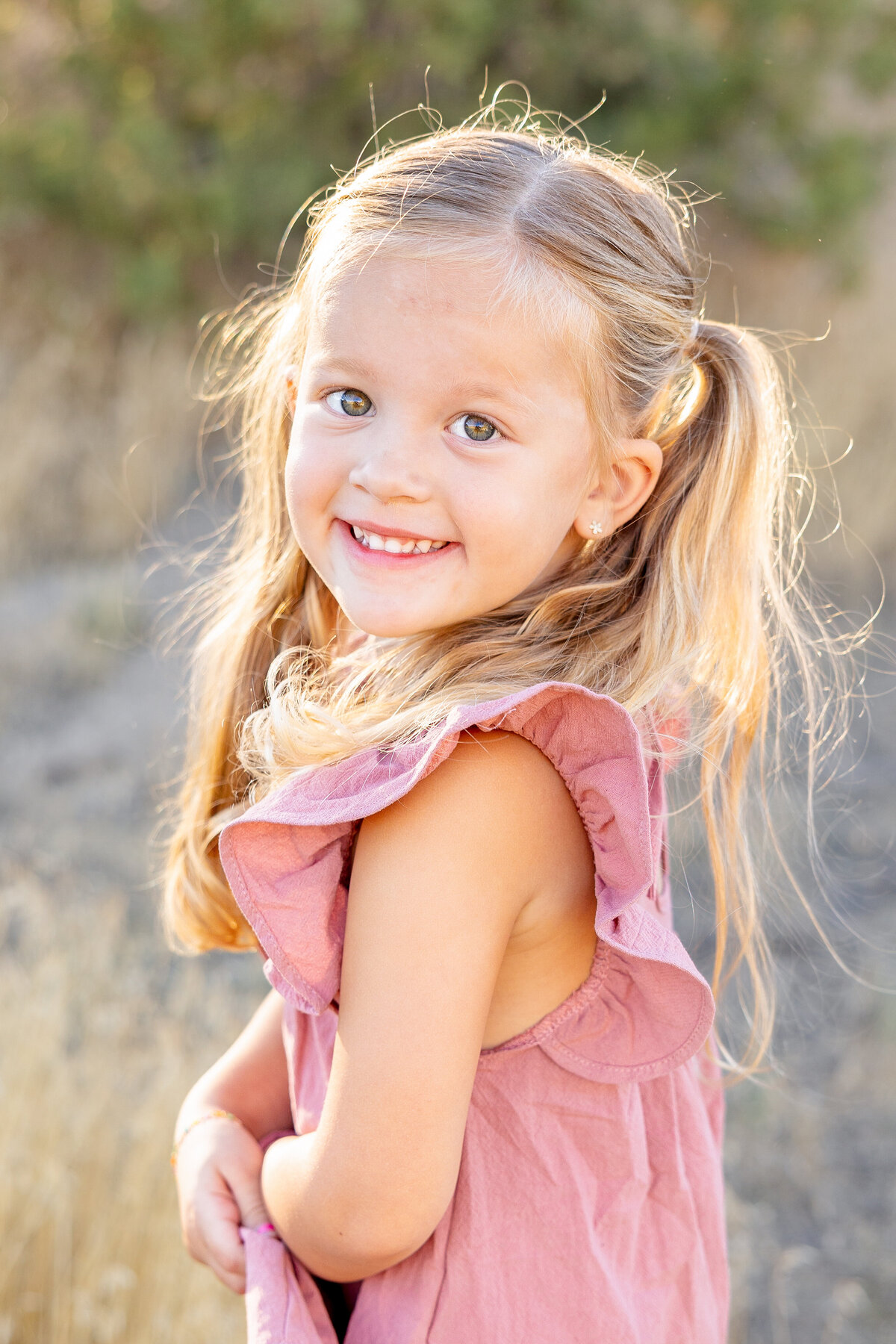 A young girl with pigtails and a pink dress smiles brightly in the golden sunlight. Her eyes sparkle with joy, and the soft, rustic background highlights her playful energy