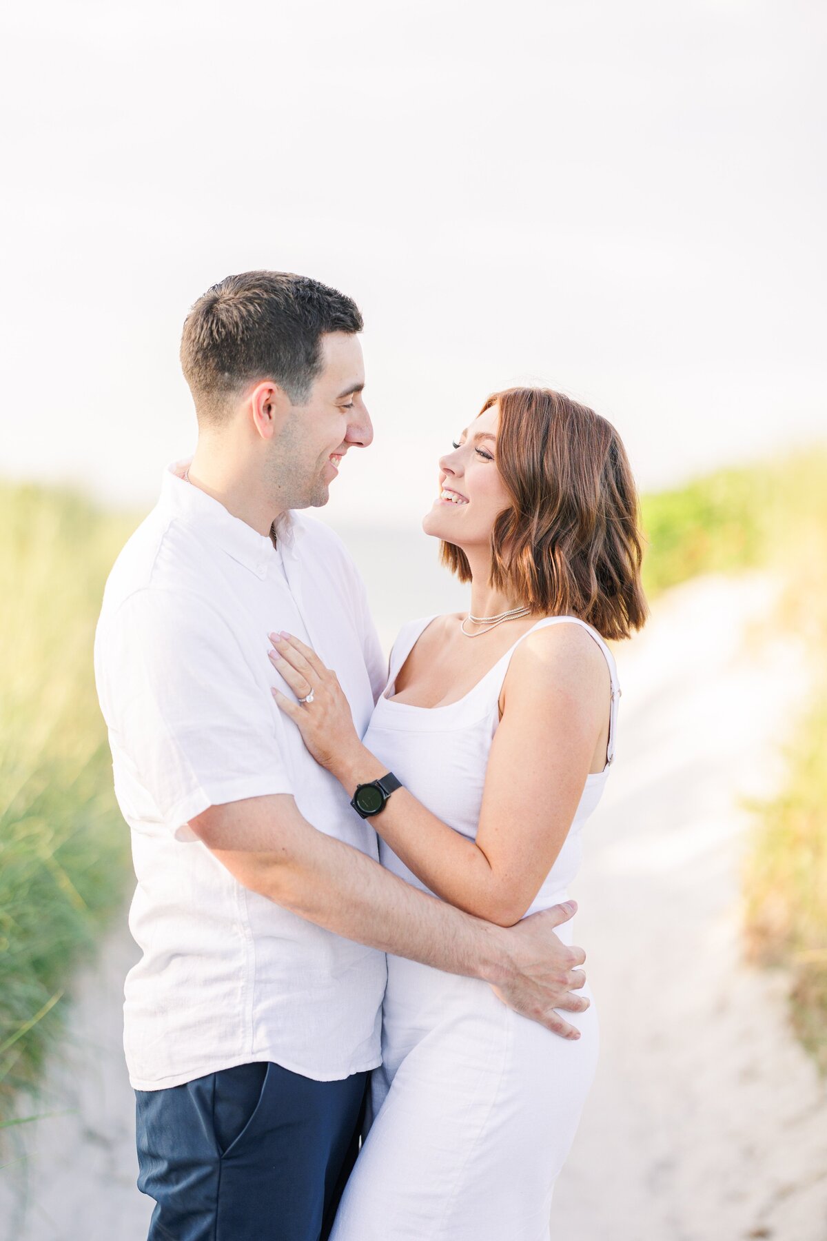 A couple hugging and laughing on a beach path at Wingaersheek Beach representing a Boston engagement photographer's portfolio.