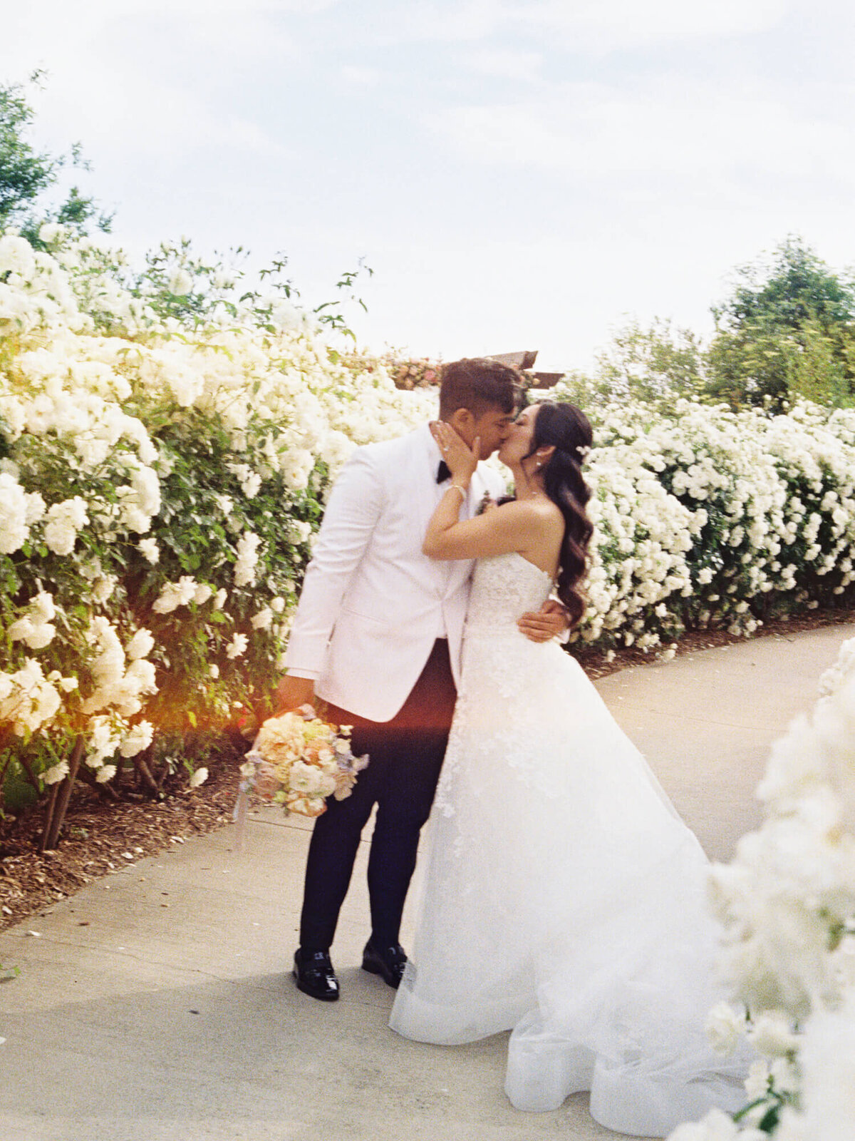 Couple kissing on a path lined with blooming white flowers. The bride wears a white gown, the groom a white suit, conveying romance and joy.