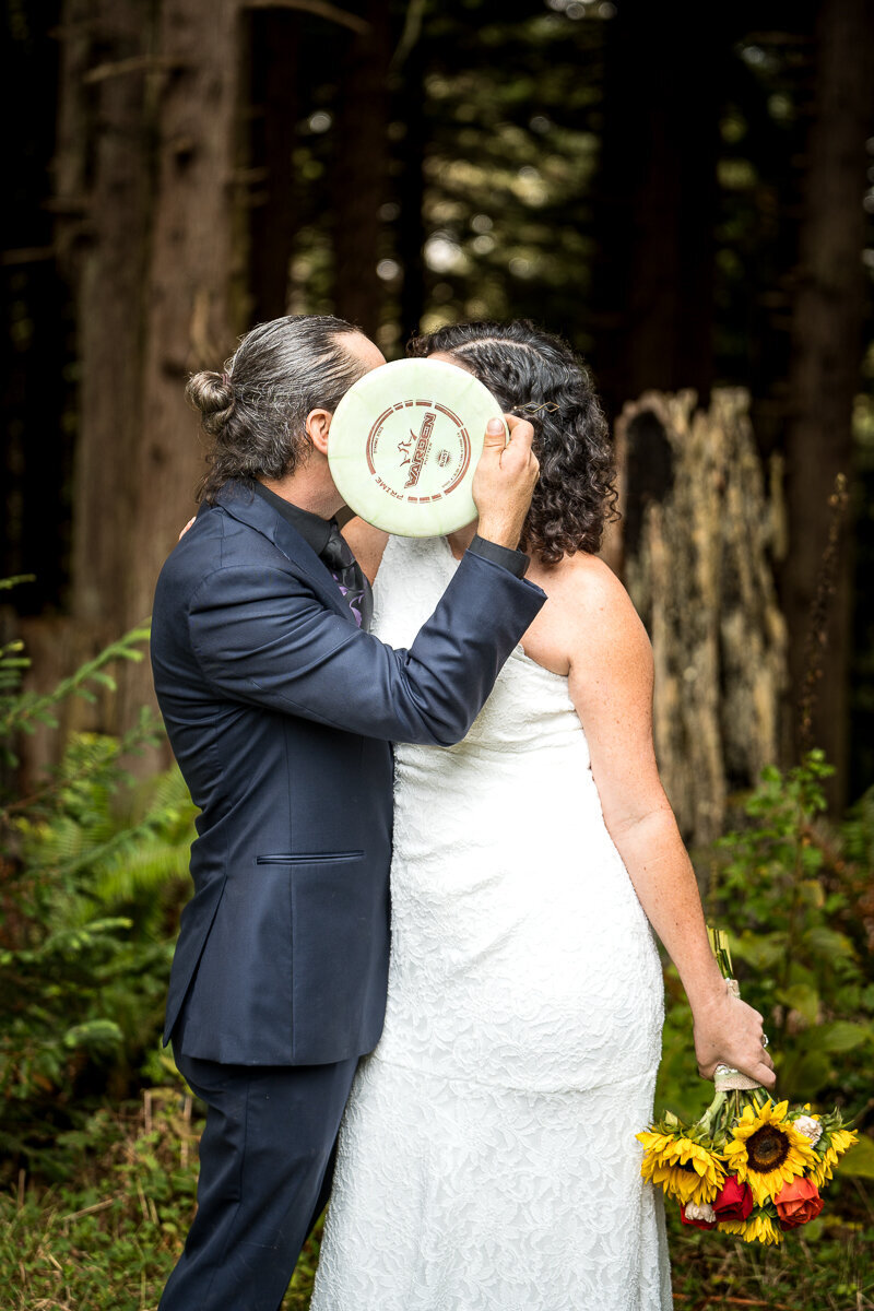 A fun and unique wedding photo of a bride and groom sharing a kiss behind a disc golf disc held by the groom, celebrating their love and passion for the sport in Arcata, CA.