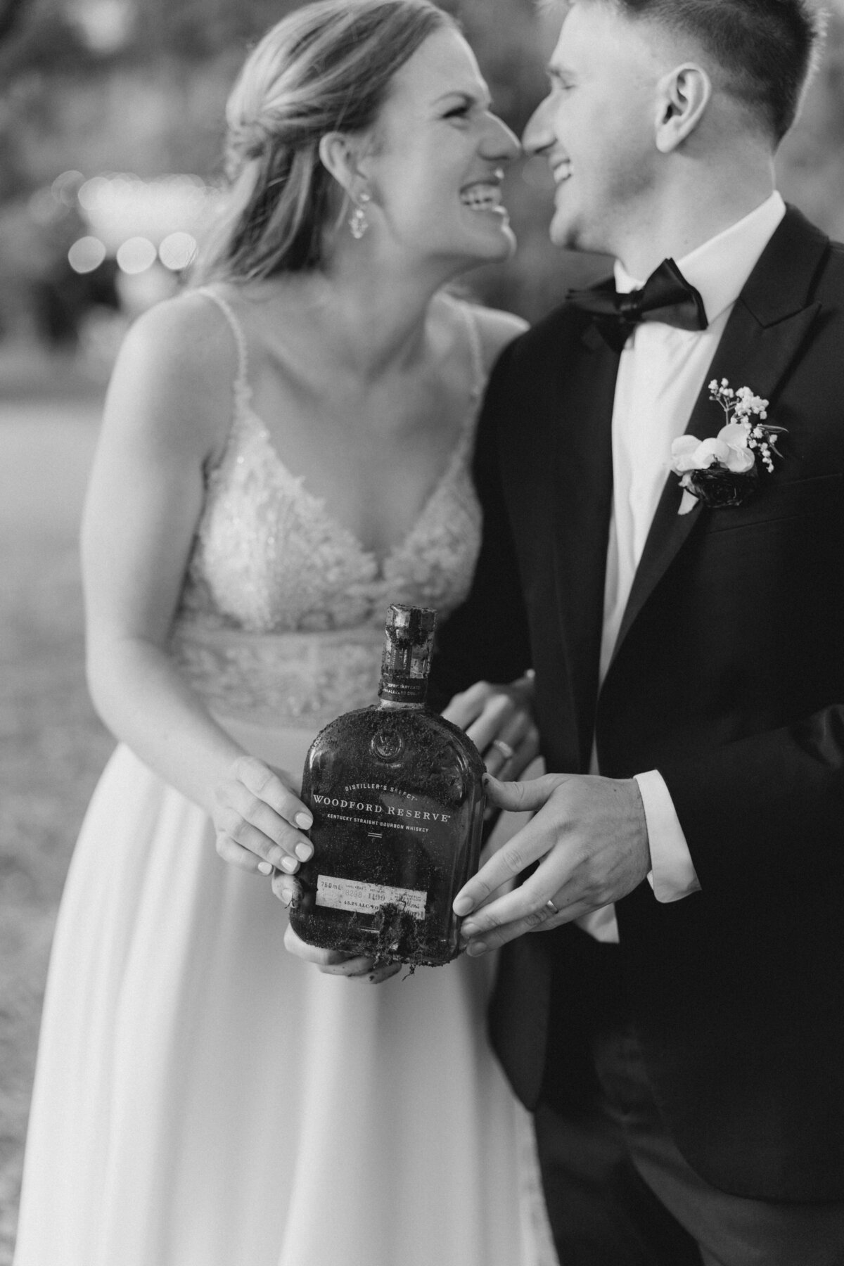 Southern tradition of burying the bourbon brings good weather on your wedding day. Bride and groom hold dirt covered bottle of bourbon and smile at each other. Charleston based black and white wedding photographer.