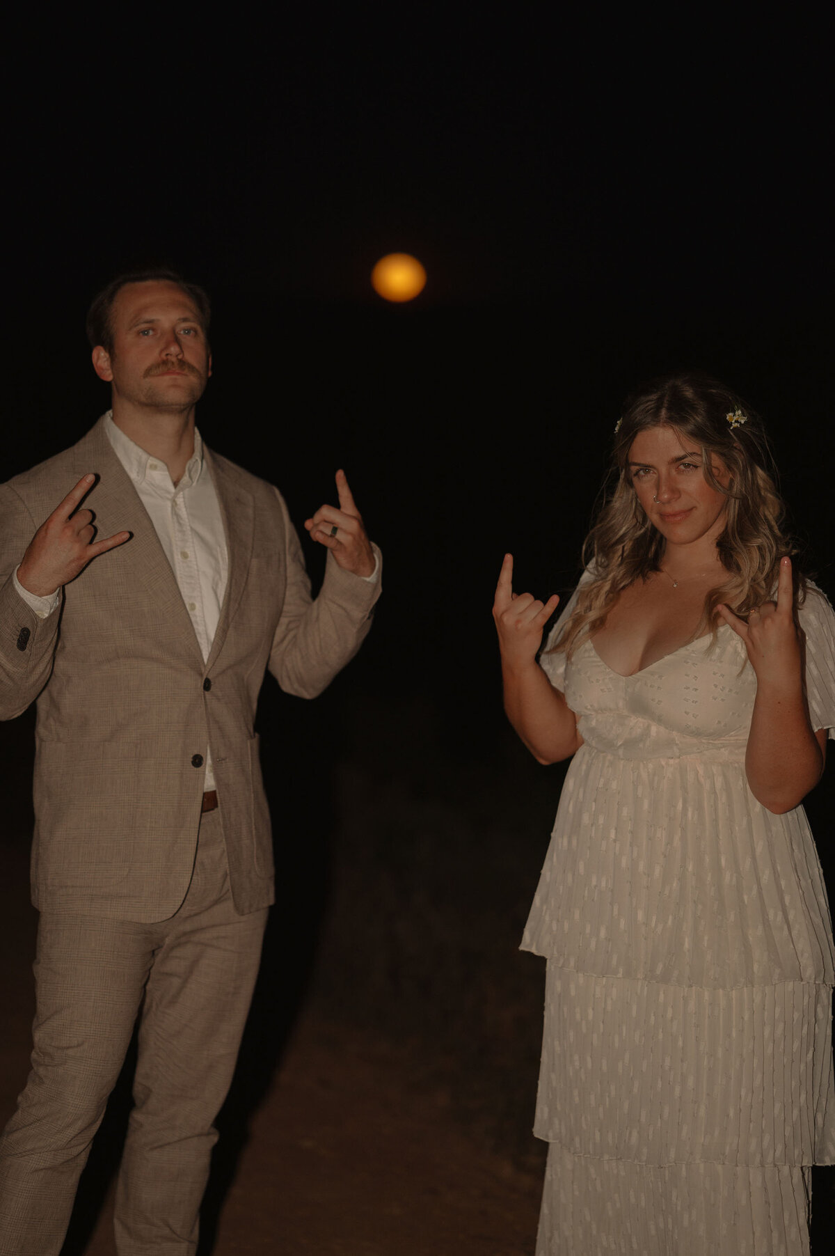 bride and groom give rock star hand signal while standing under a black sky and orange moon