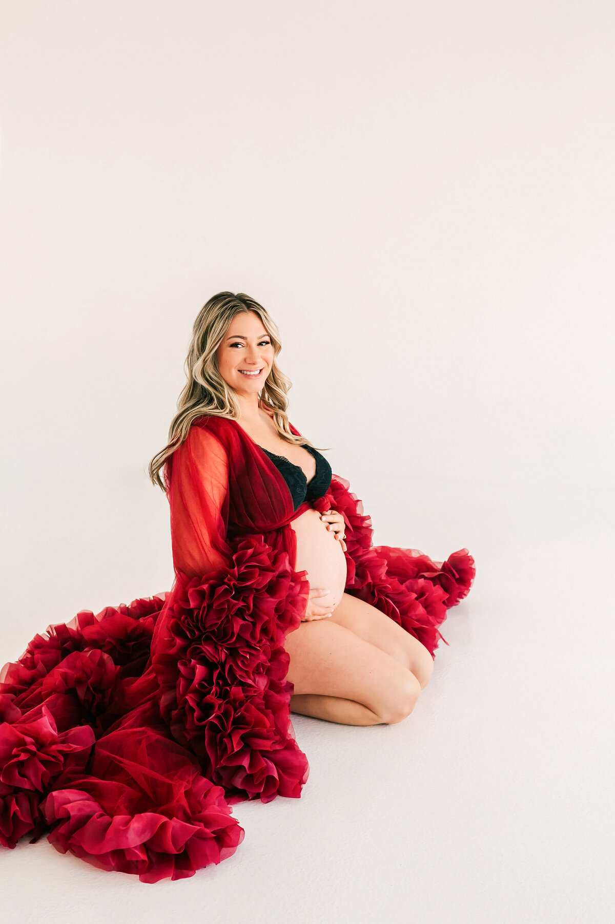 maternity photo of mom kneeling on ground smiling holding baby bump