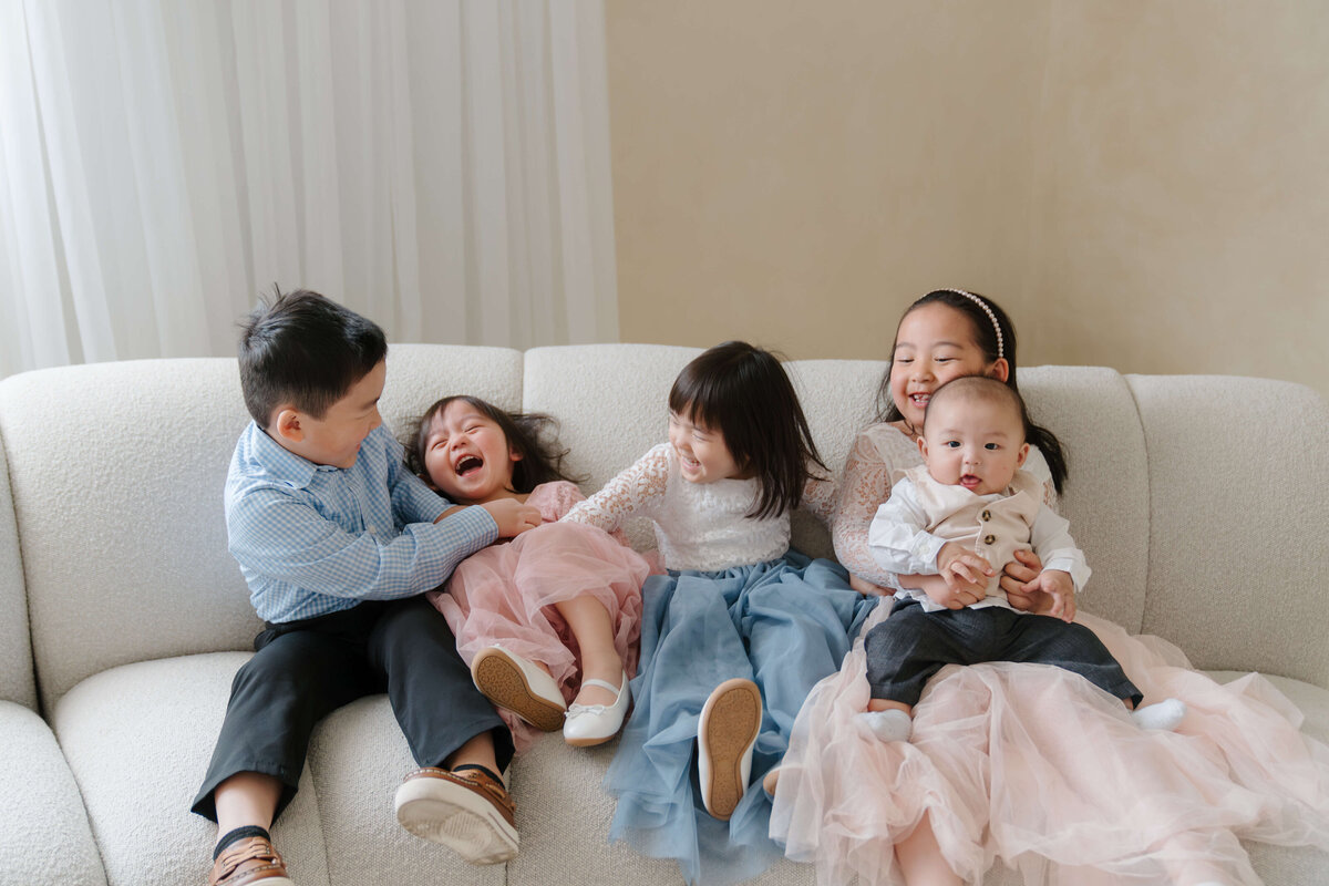 Five young siblings laughing together on a modern cream couch during a joyful family studio session.