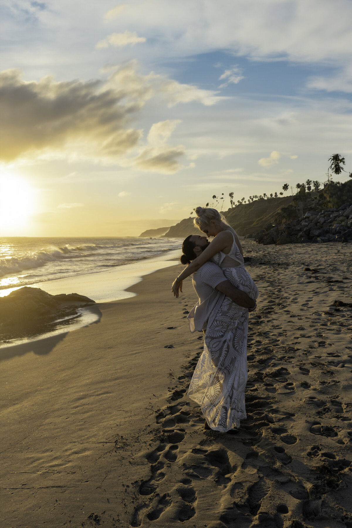 Bride and groom on the beach of Leo Carillo Malibu during sunset where the groom is picking the bride and embracing her in her lace gown