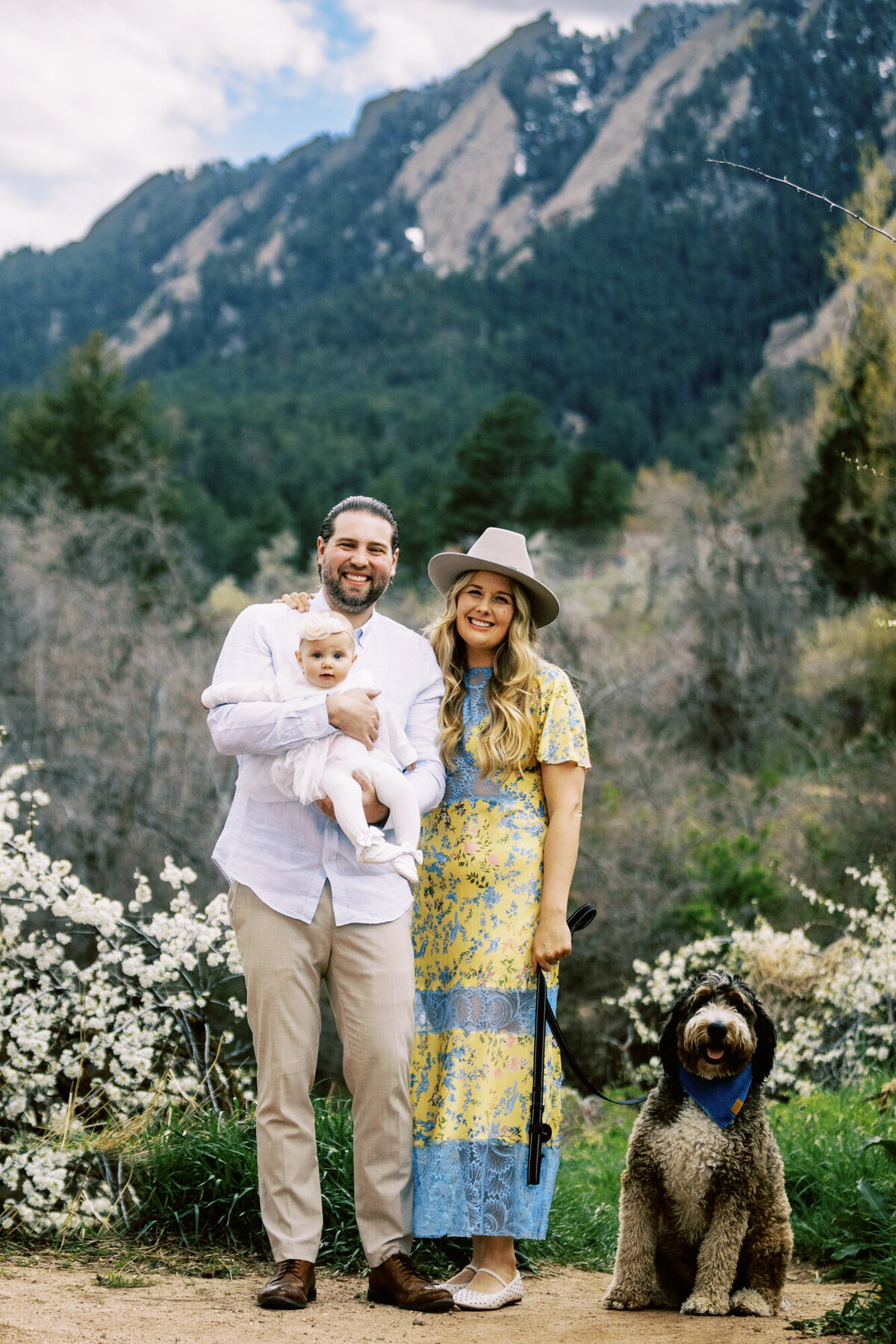 parents, their baby girl, and pet dog stand on a dirt path in boulder colorado in front of the flatiron mountains