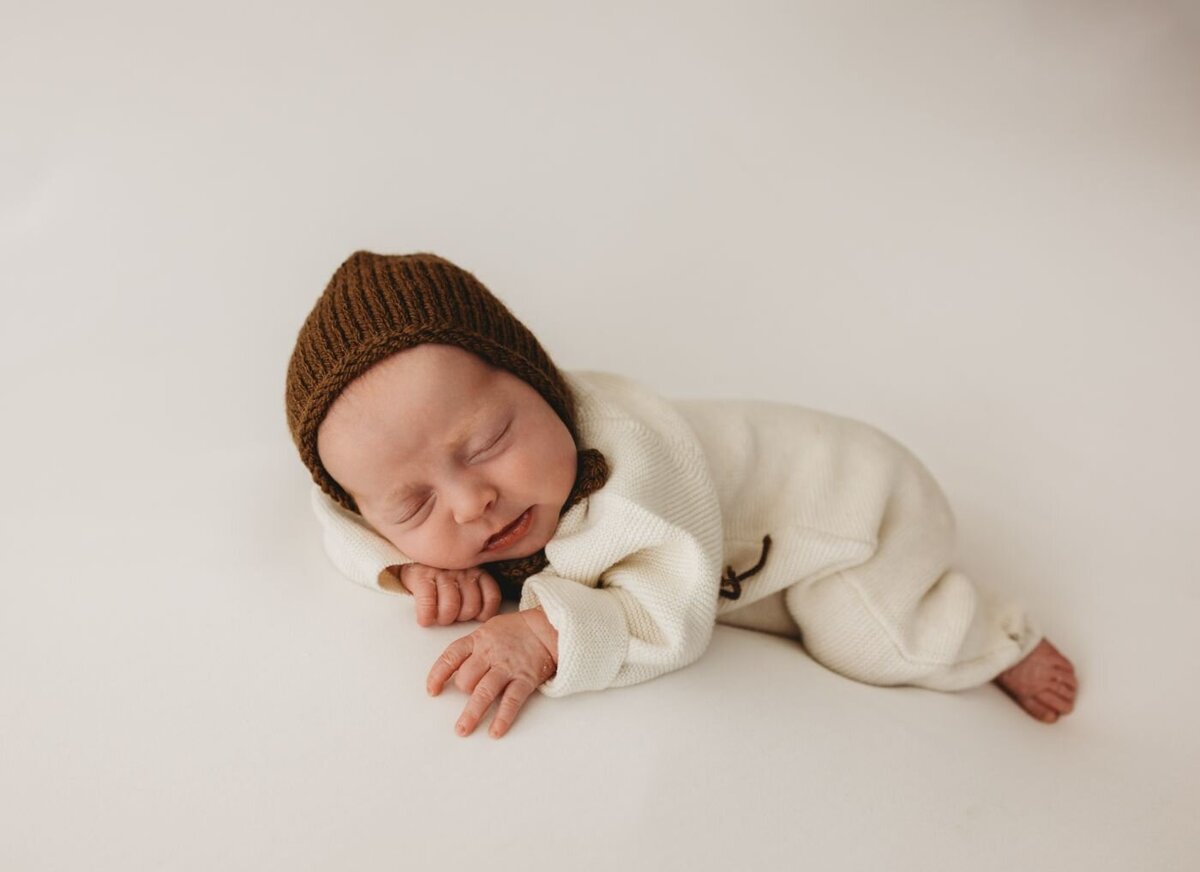 Serene studio portrait of a newborn baby sleeping soundly on a plain white surface, dressed in a soft cream romper and a brown knitted hat in UK