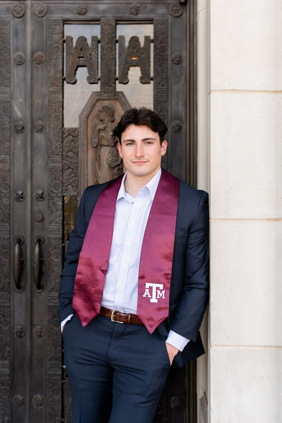 Texas A&M senior guy with a soft smile and hands in pockets leaning against front door of Administration Building with Aggie stole and formal attire