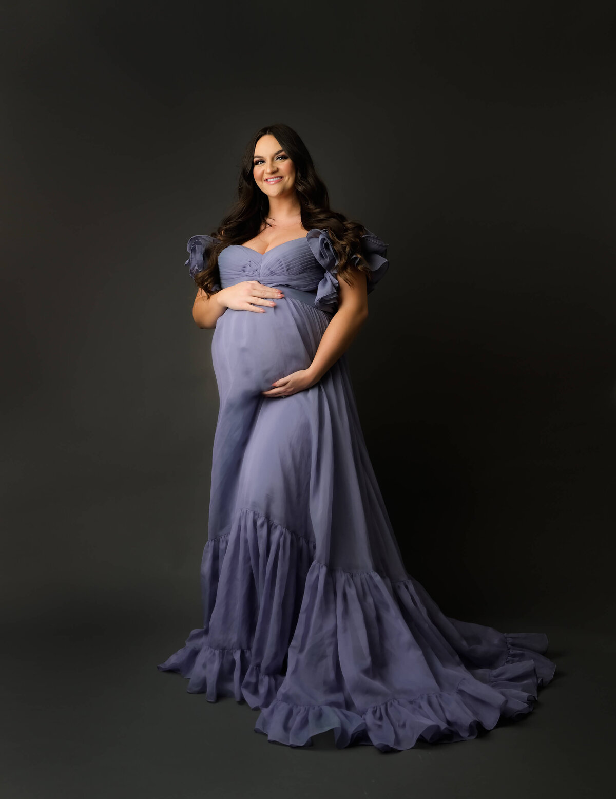 Beth Benecki of E. Benecki Photography captures an expectant mother in a purple chiffon gown  for this maternity photo session against a dark gray background.