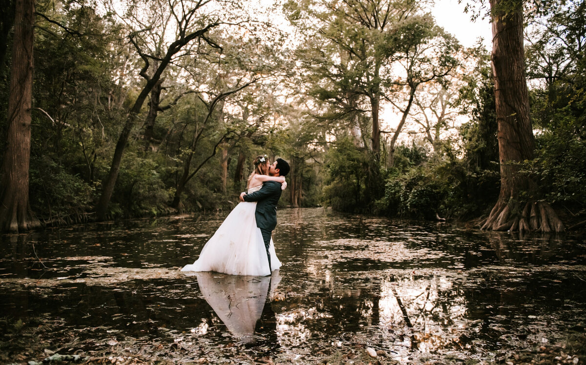 Couples Photography, Man in blue suit and woman in a wedding dress hold each other closely in the middle of a deep creek with trees surrounding them on all sides.