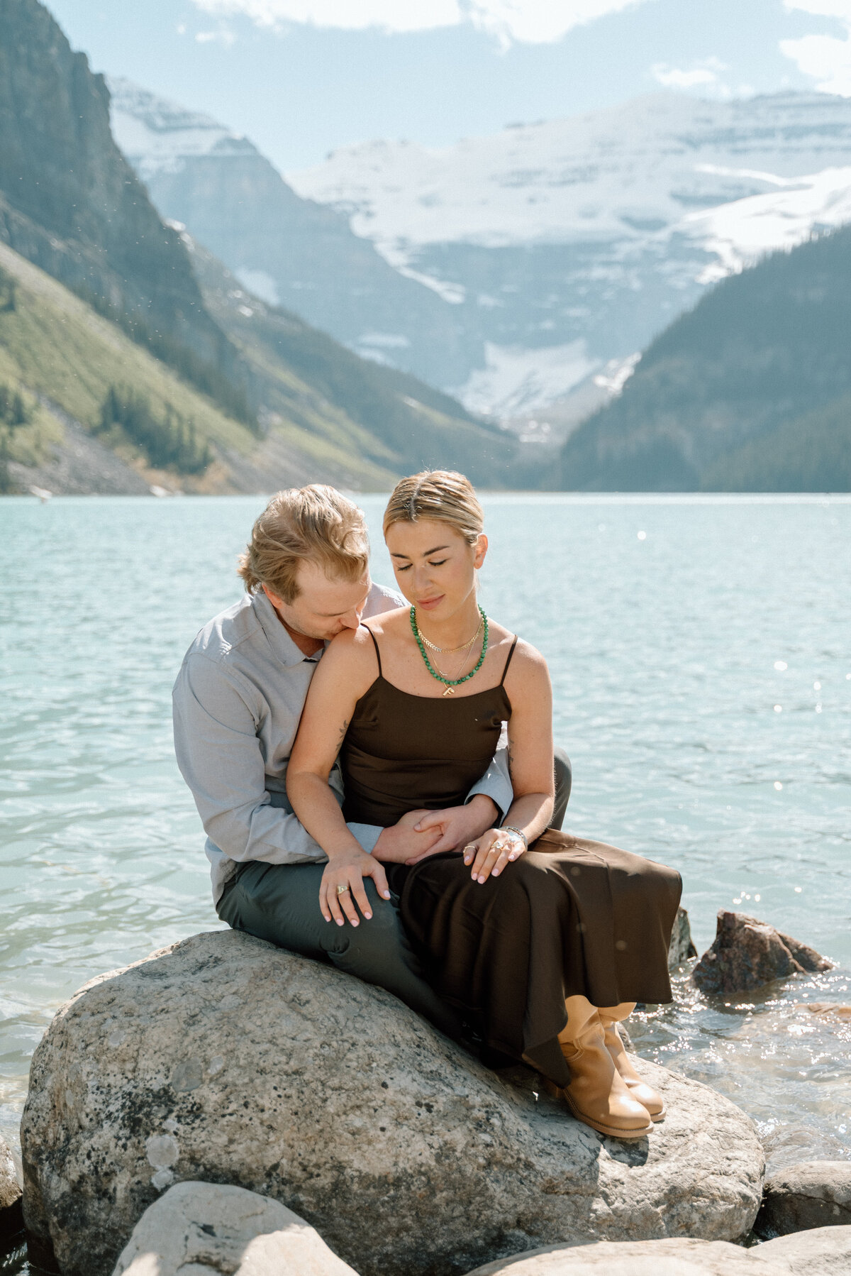 Engagement photo with couple seated on large rock by Lake Louise shoreline.