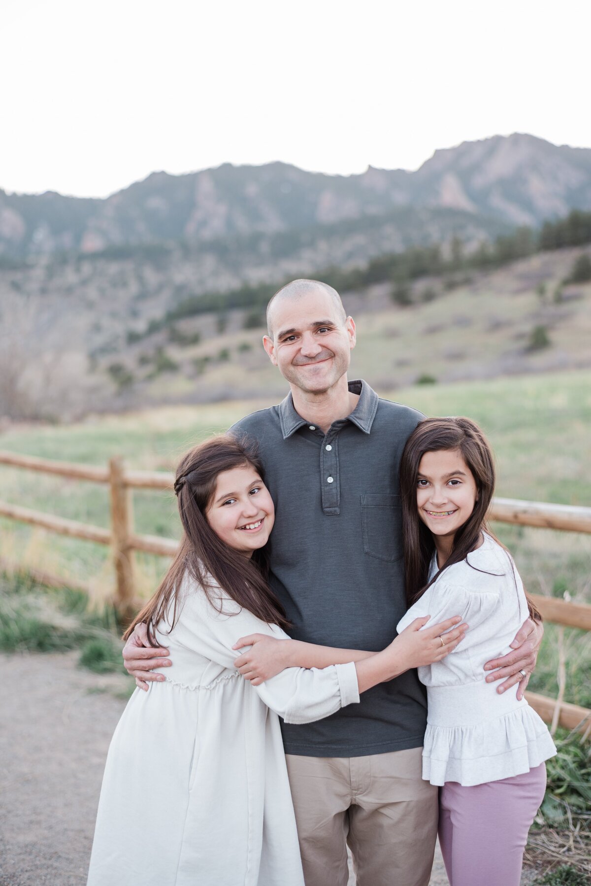 A dad smiles for the camera while his two young daughters hug him and they all look at denver family photographer