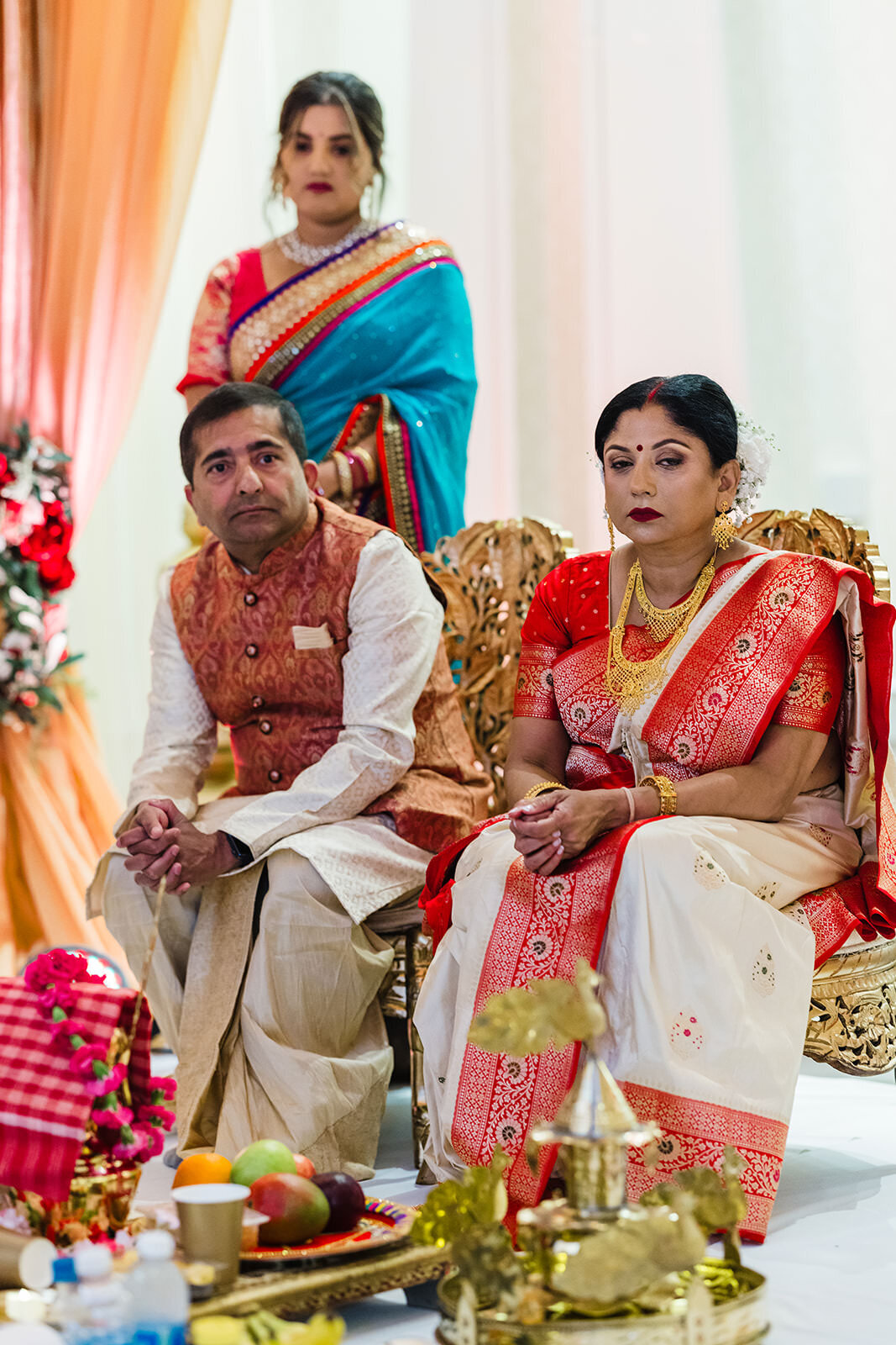 Two seated adults, a man in traditional Indian attire and a woman in a red and white saree, appear focused on a ceremony, with a standing woman in the background.