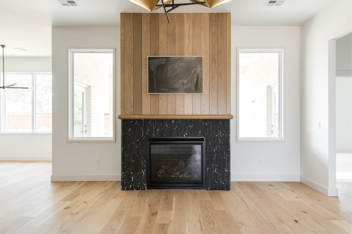 Modern living room with white oak floors, black marble fireplace surround, vertical wood paneling, and sculptural brass chandelier framed by natural light from two large windows.